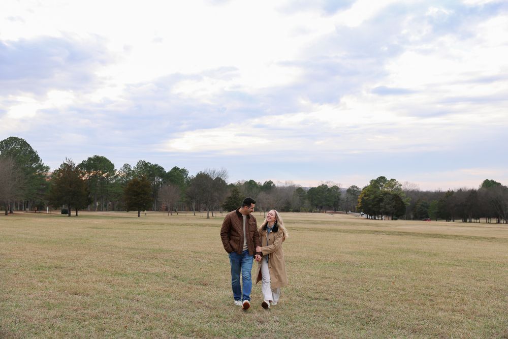 A couple walks together across a vast grassy field on a cloudy day.