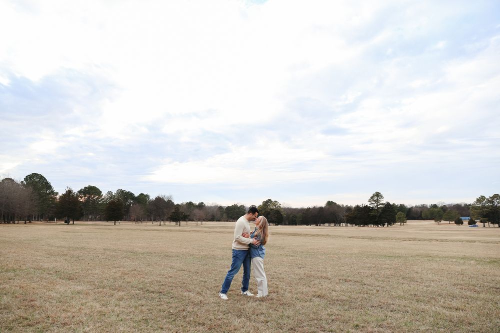 A couple embracing in an open grassy field under a cloudy sky.