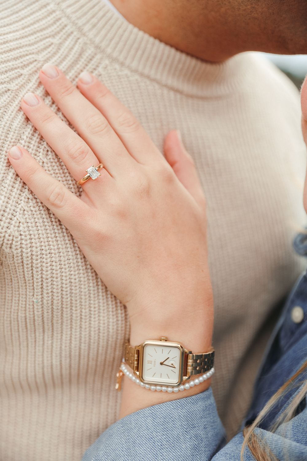 Close-up view of a delicate gold watch and ring on a person's hand wearing a cream-colored knit sweater.