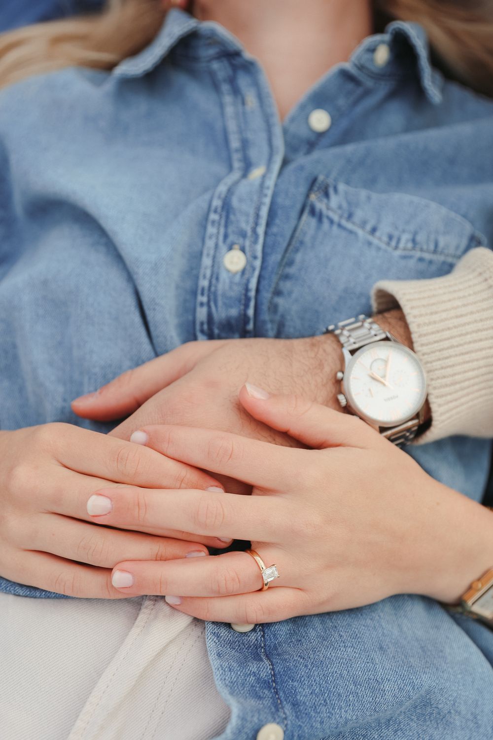 Close up of hands wearing engagement ring and watch against denim background showing romantic proposal moment.