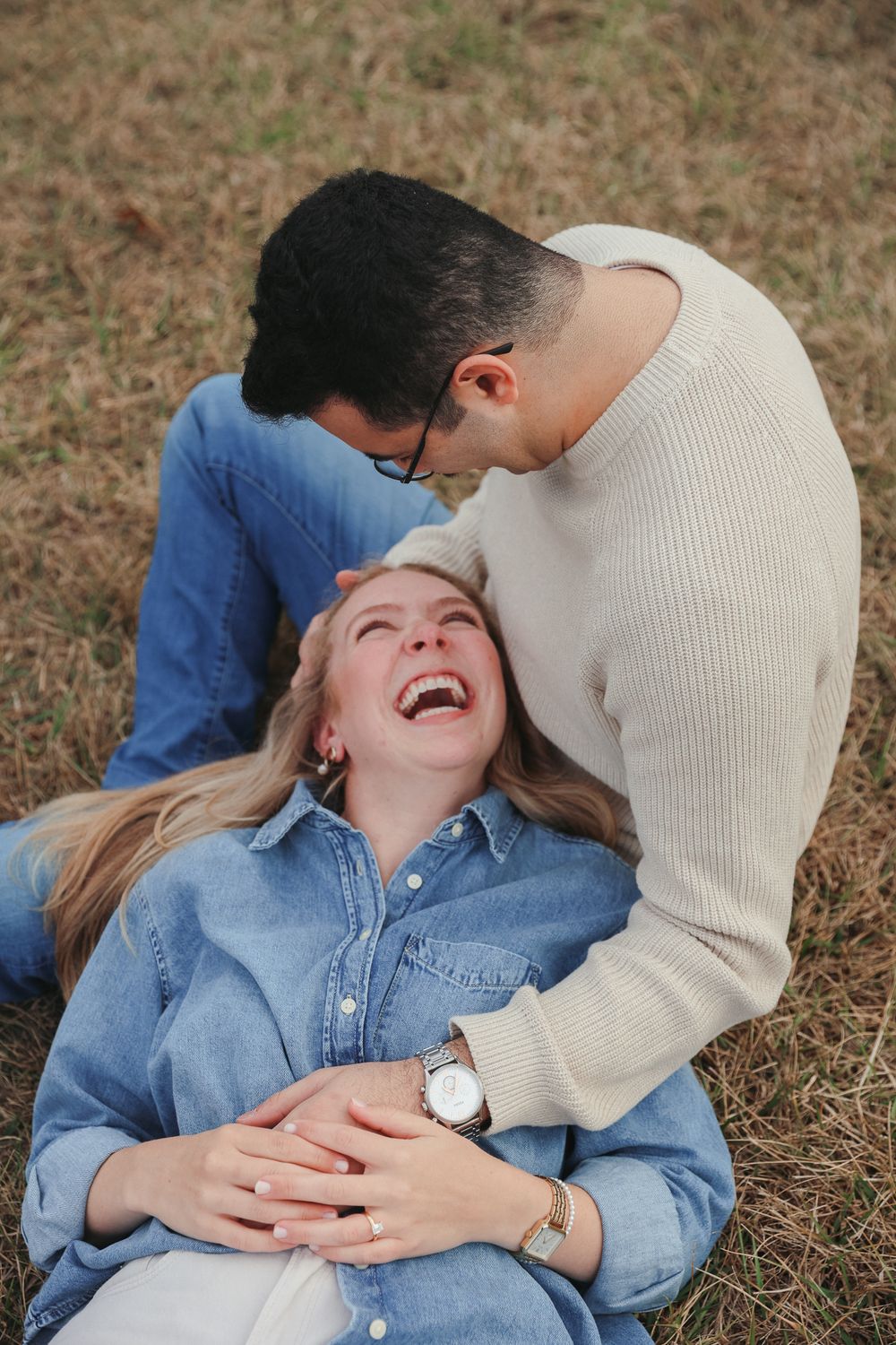 A couple lying in dry grass sharing a joyful, intimate moment together in casual denim and cream attire.