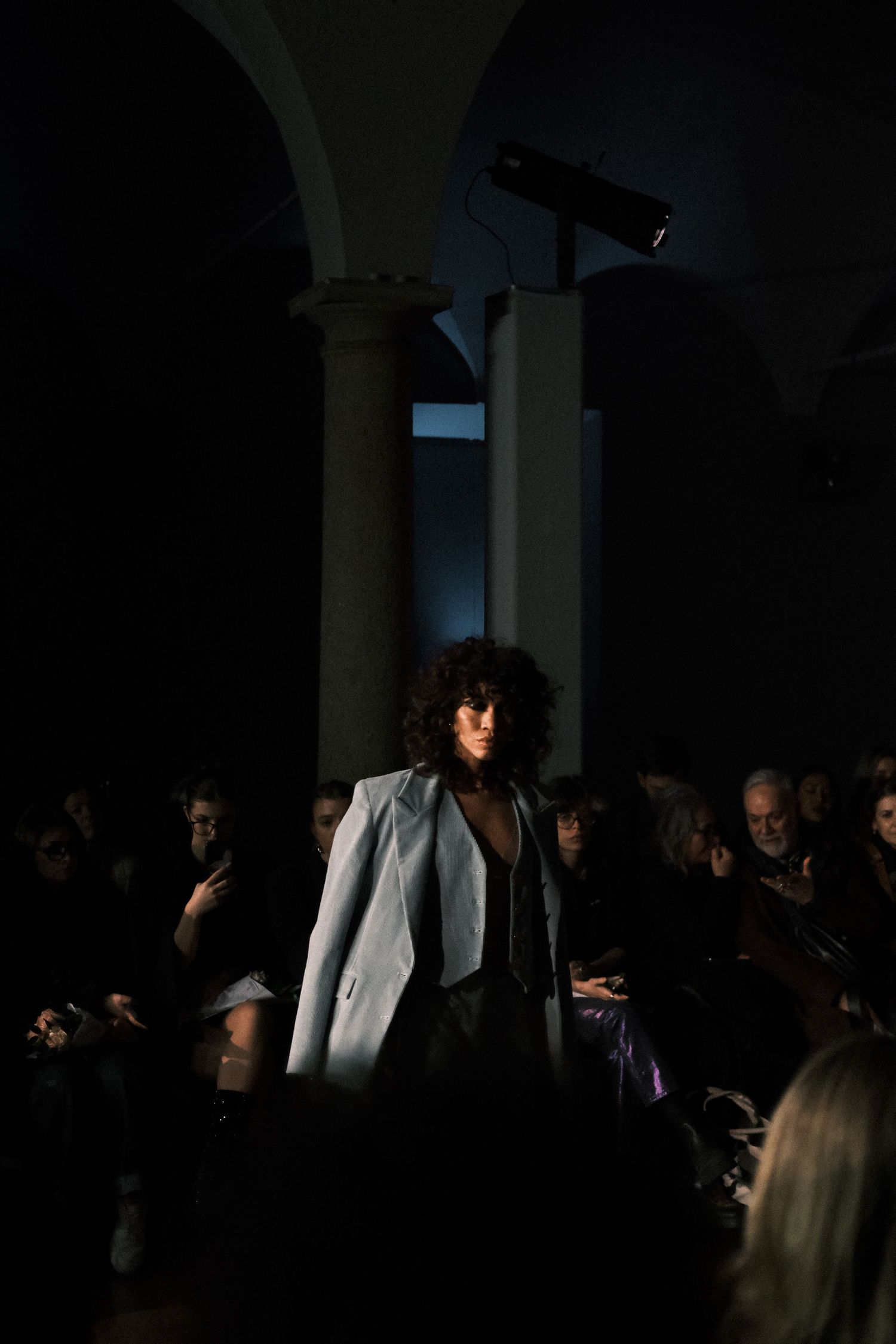 A fashion show audience sits in darkness as models walk the runway in dramatic lighting and white outfits.