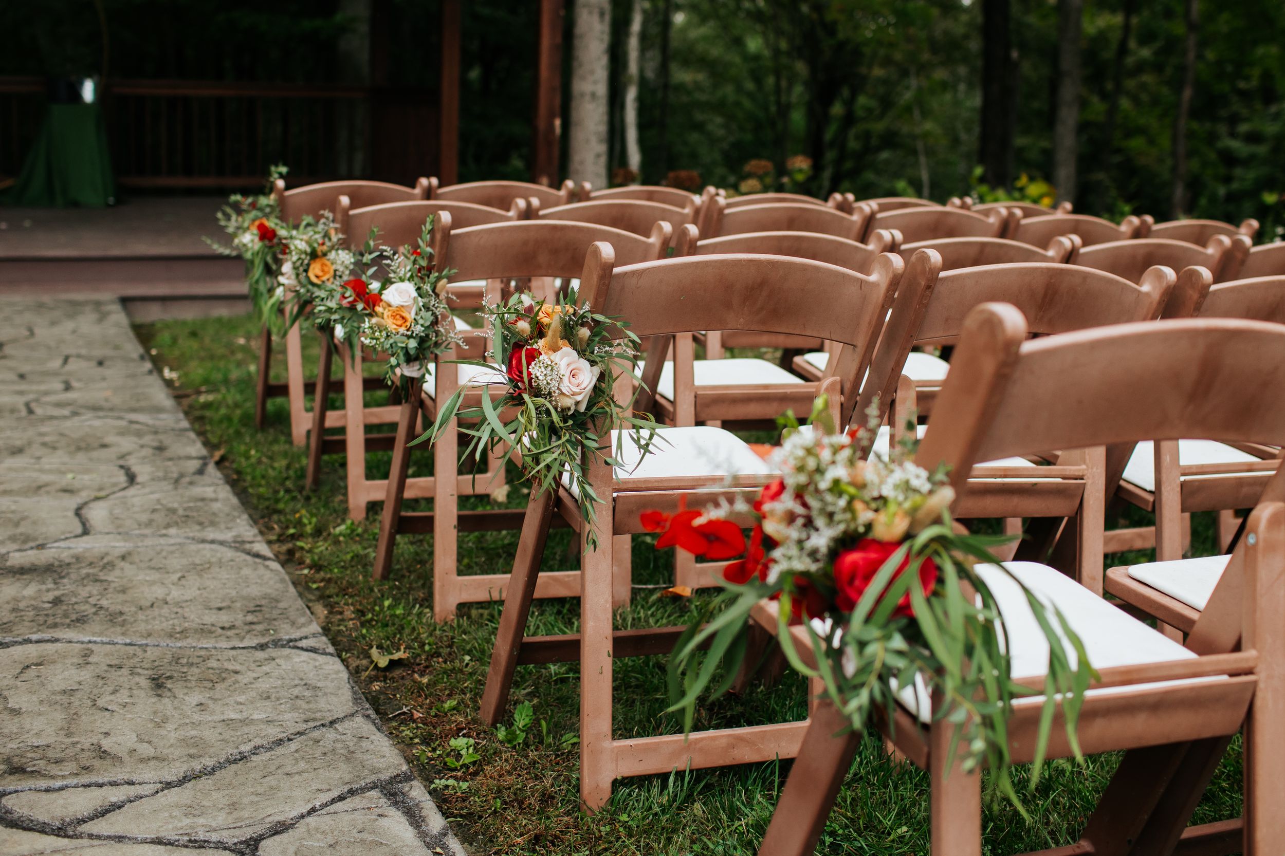 Brown wooden folding chairs decorated with floral arrangements line a stone pathway for an outdoor wedding ceremony.