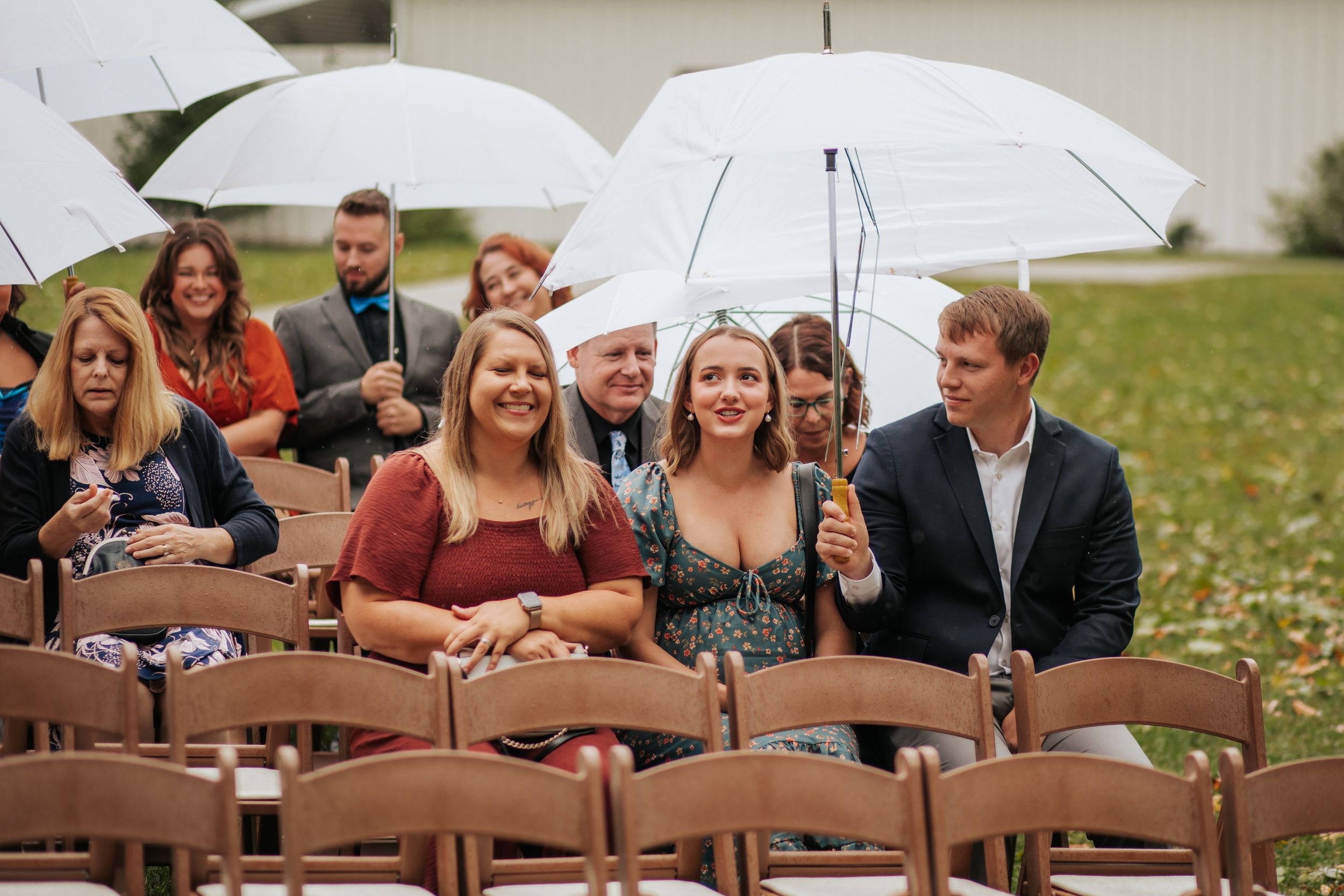 Wedding guests laugh and smile while sitting in wooden chairs under white umbrellas during an outdoor ceremony.