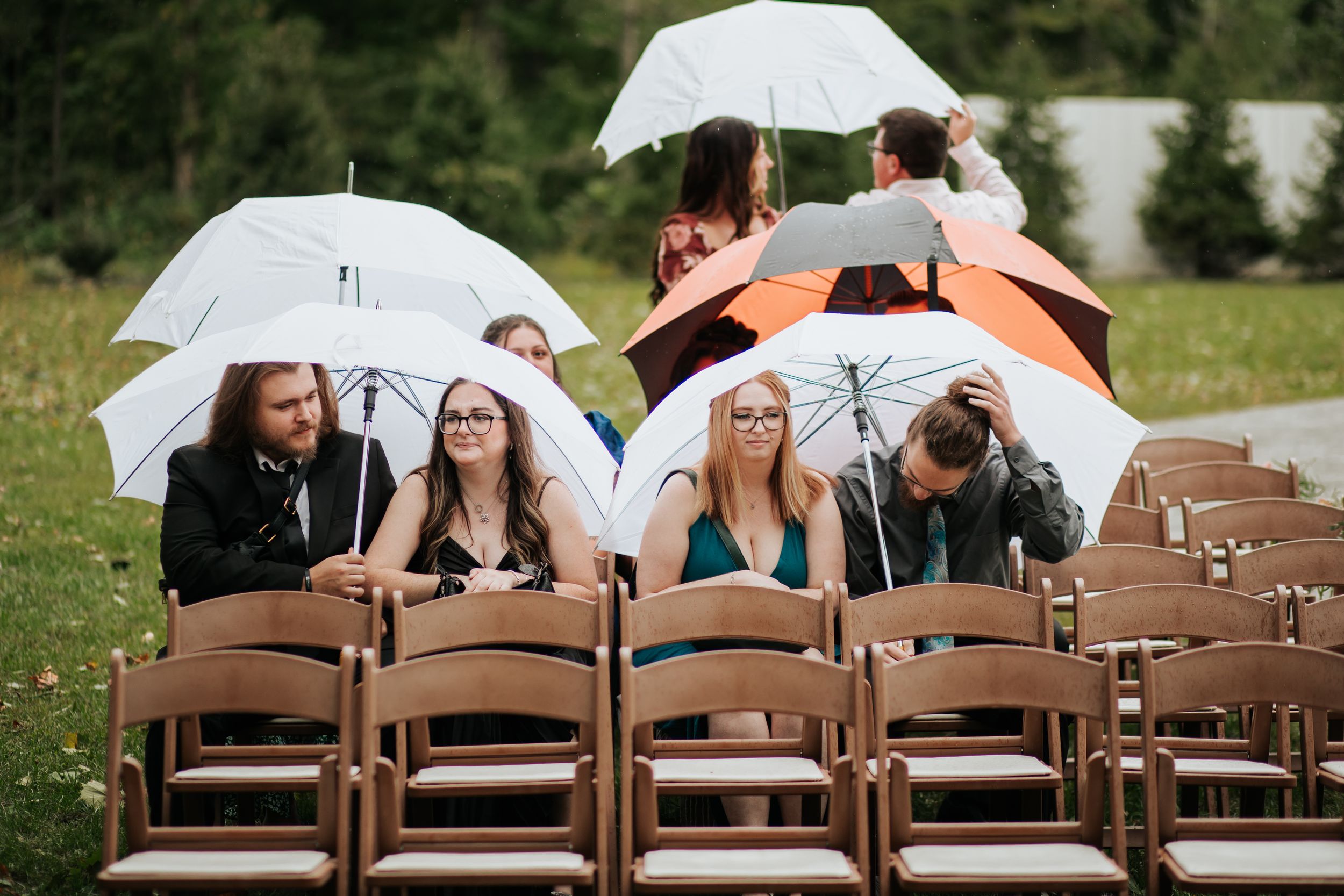 Wedding guests seated in brown chairs holding white umbrellas during an outdoor ceremony.