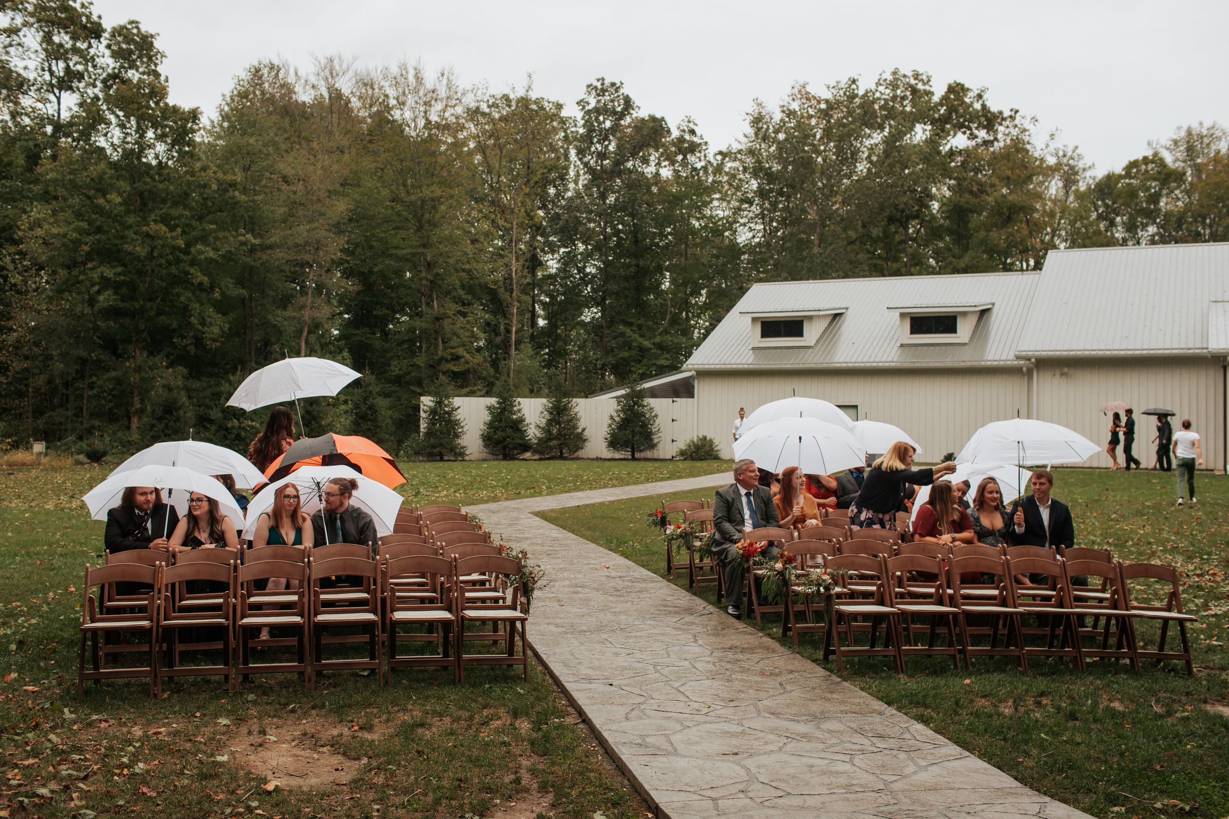 Outdoor wedding ceremony setup with wooden chairs and white umbrellas on a concrete path leading to a white barn.