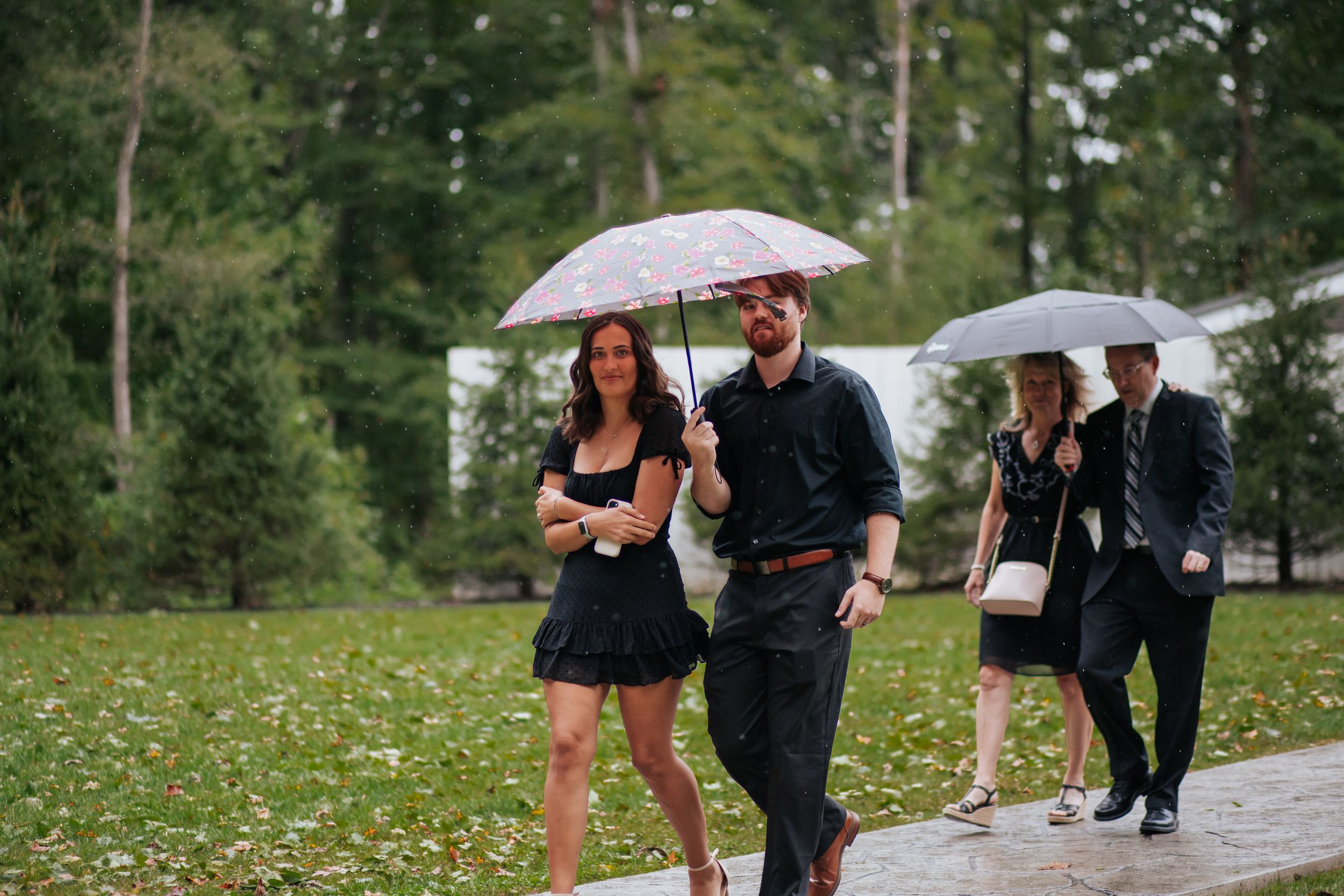 Wedding party walks down a path under umbrellas on a rainy day surrounded by greenery and trees.