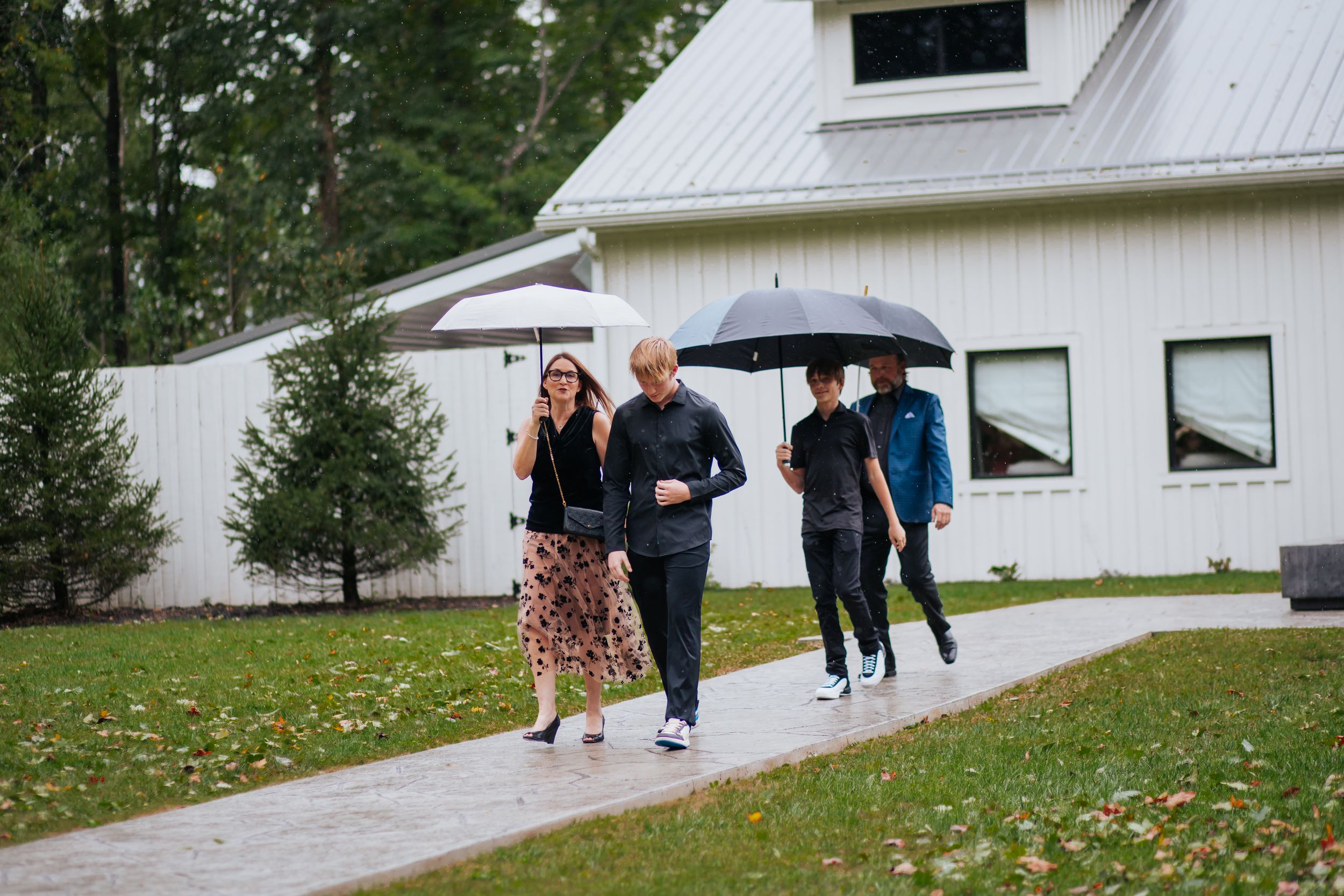 People walking together with umbrellas on a rainy day outside a white barn-style building with autumn leaves on the ground.