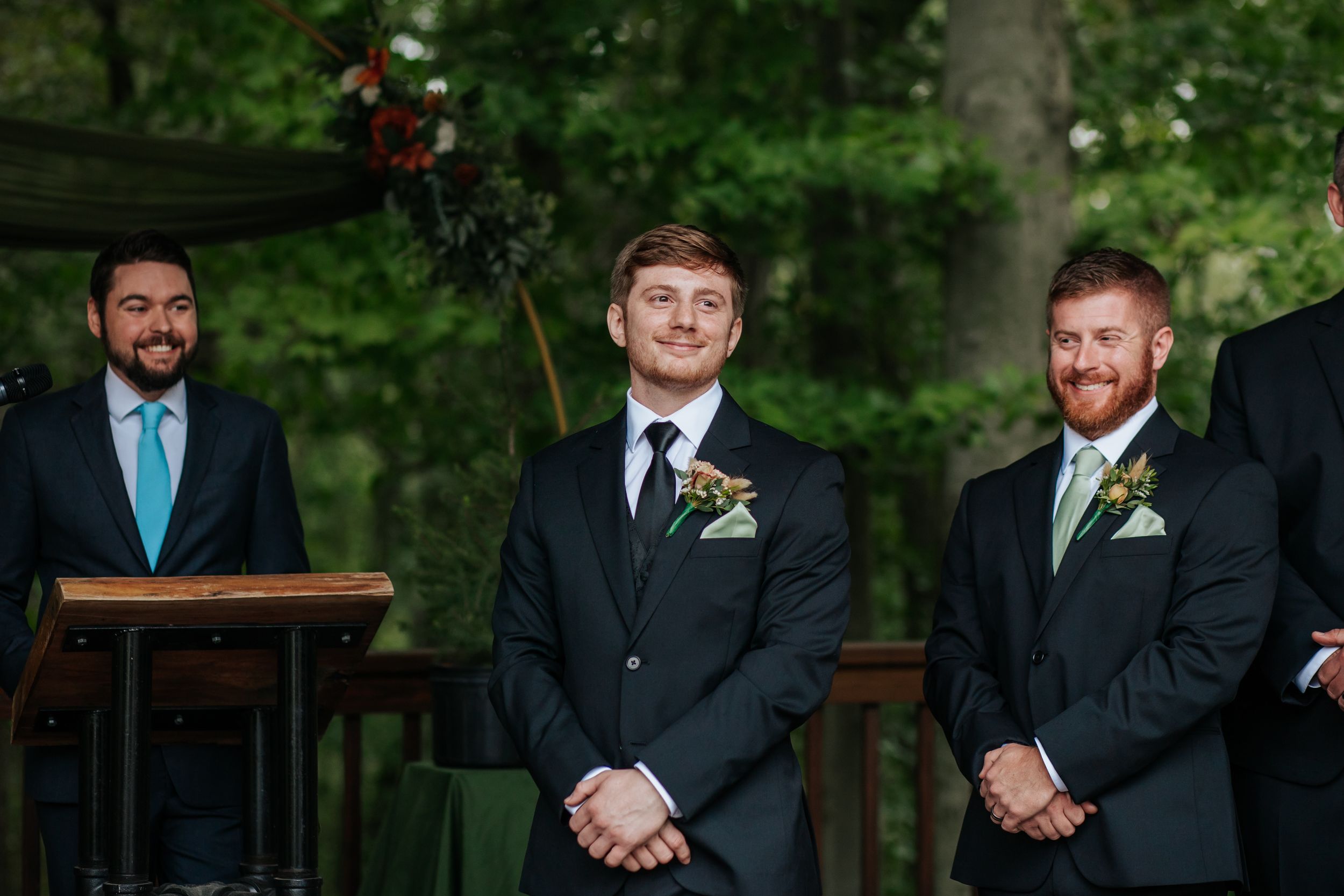 Groomsmen in dark suits with sage green accents stand in line at an outdoor wedding venue.