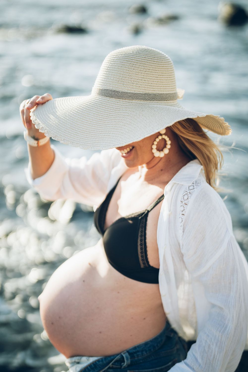 Pregnant woman in white sun hat poses by ocean wearing black swimsuit.