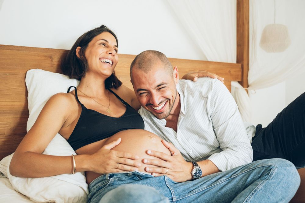 A cheerful couple relaxes on bed together while touching a pregnant belly in an intimate maternity moment.