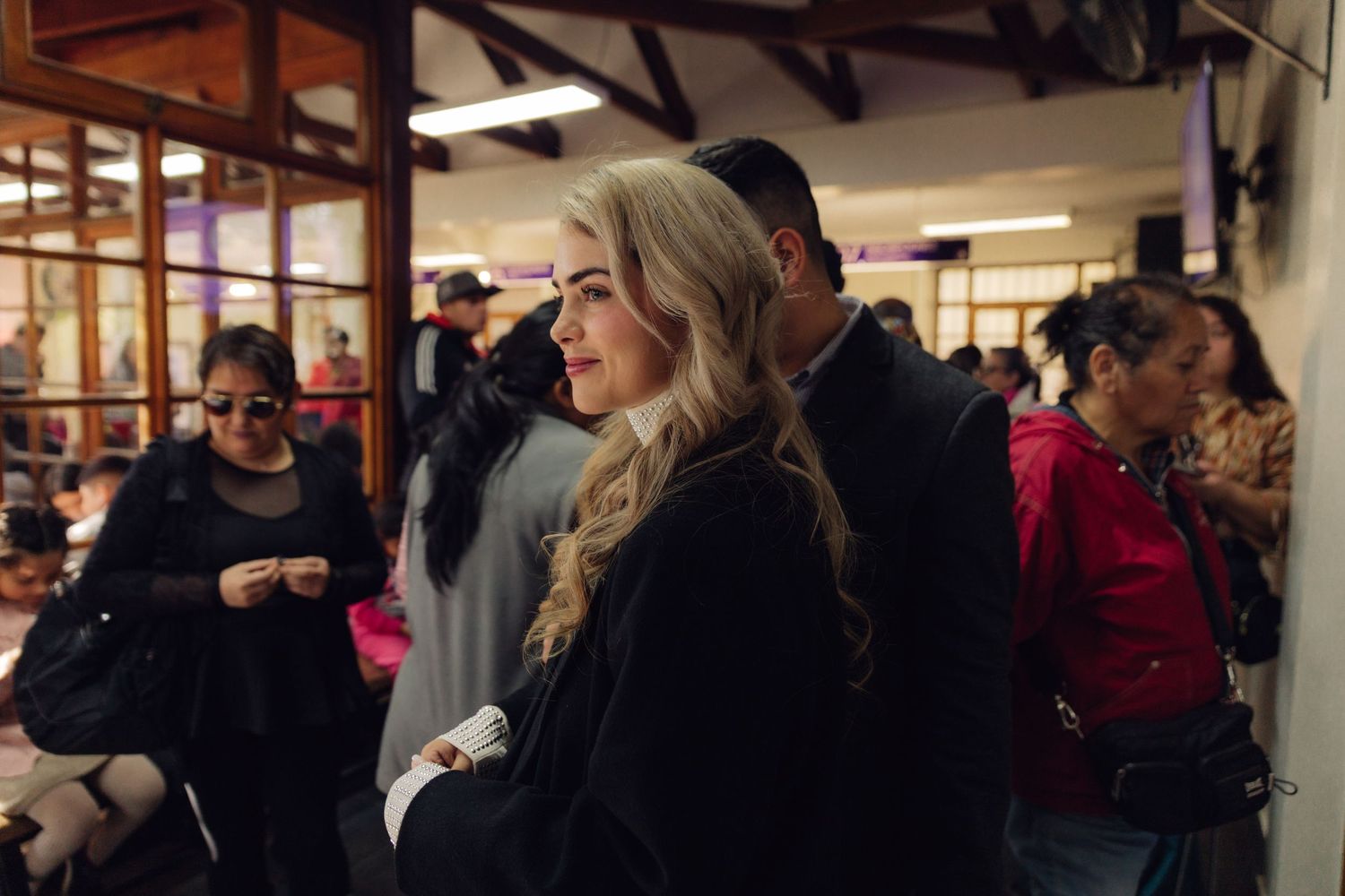 Blonde woman in black coat standing in crowded indoor venue, looking elegant at a formal gathering.