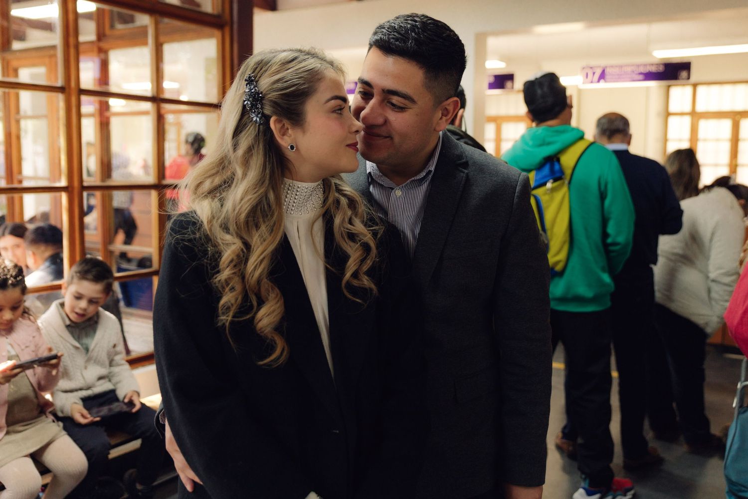 Couple sharing a kiss indoors at a civil ceremony, surrounded by guests in a bright public building.