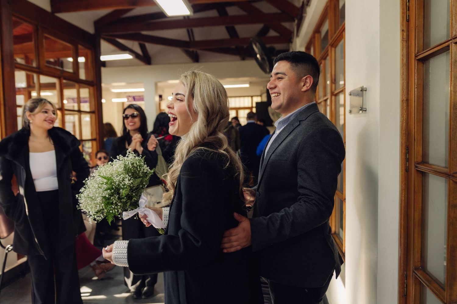 Joyful couple laughing together indoors, bride holding white bouquet with wedding guests in background.