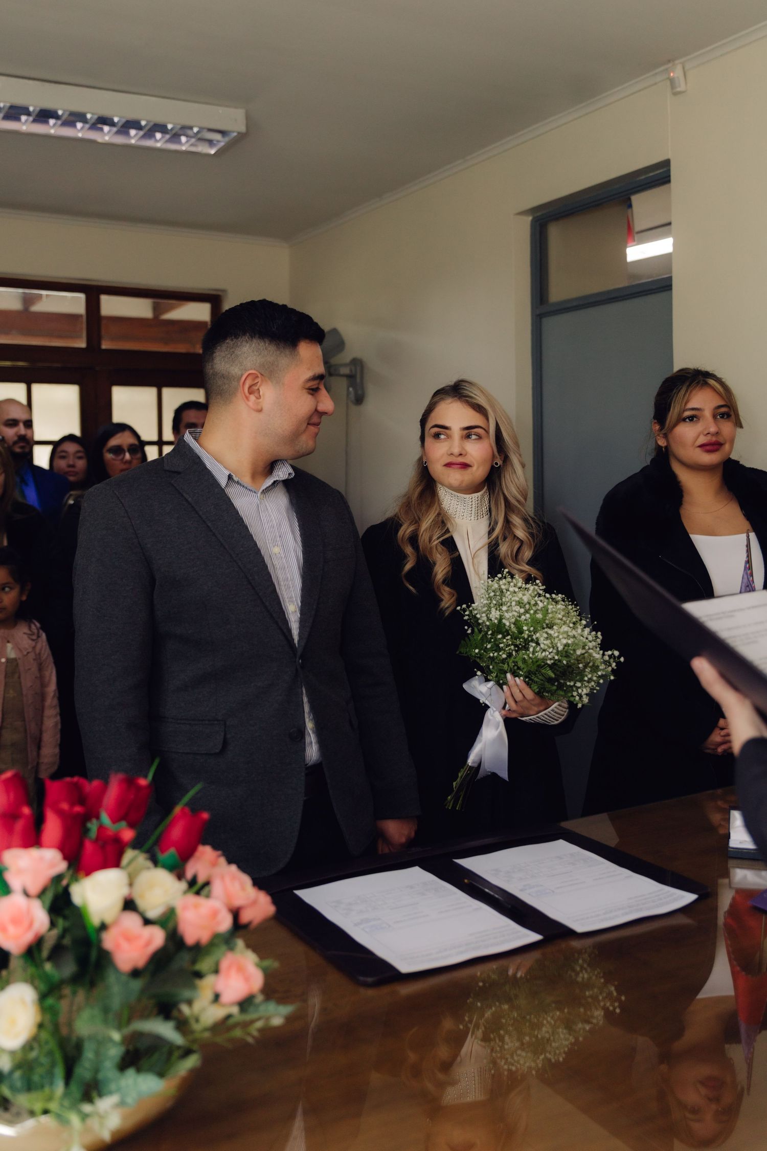 Couple at civil wedding ceremony with signed documents and flower arrangements on desk.