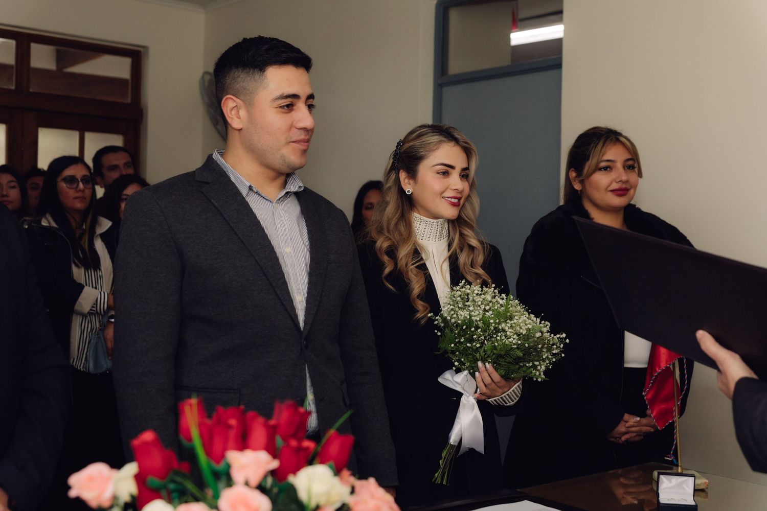 Smiling couple holding hands at civil wedding ceremony with floral arrangements present.
