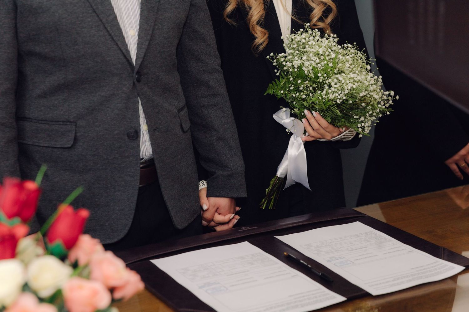 Close-up of couple holding hands with bouquet at civil wedding signing ceremony.