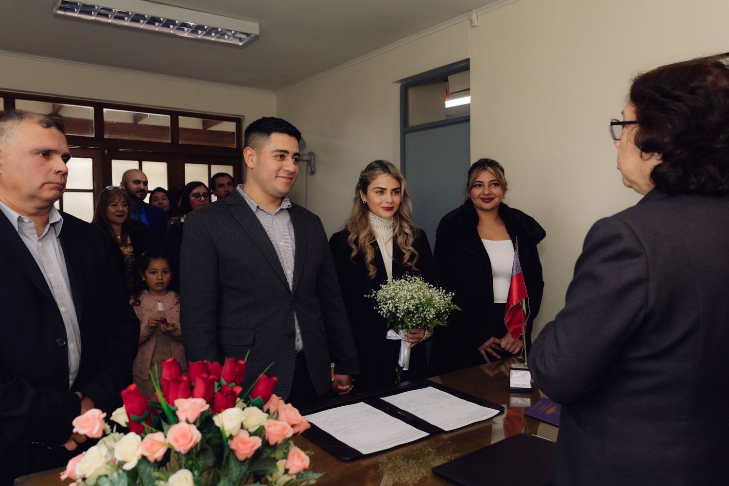 Couple stands before officiant during civil wedding ceremony with guests and floral arrangements in background.