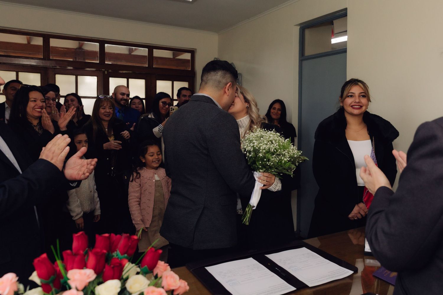 Newlyweds share first kiss at civil wedding ceremony as guests applaud and bridesmaid smiles nearby.