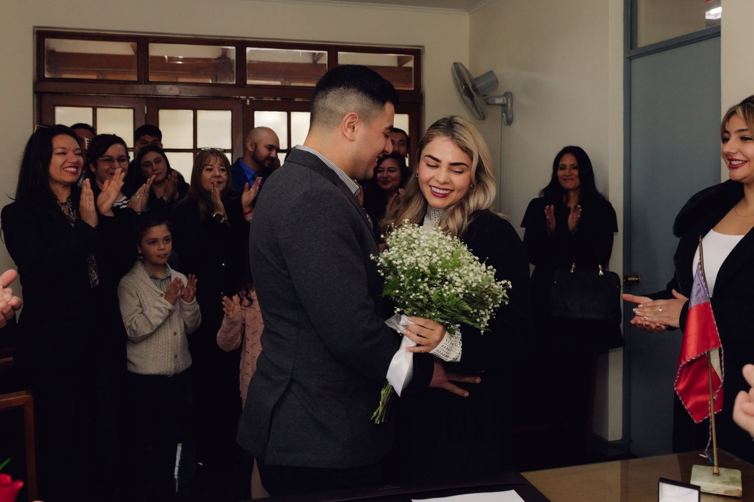 Groom presents bride with white bouquet as wedding guests applaud during civil marriage celebration indoors.