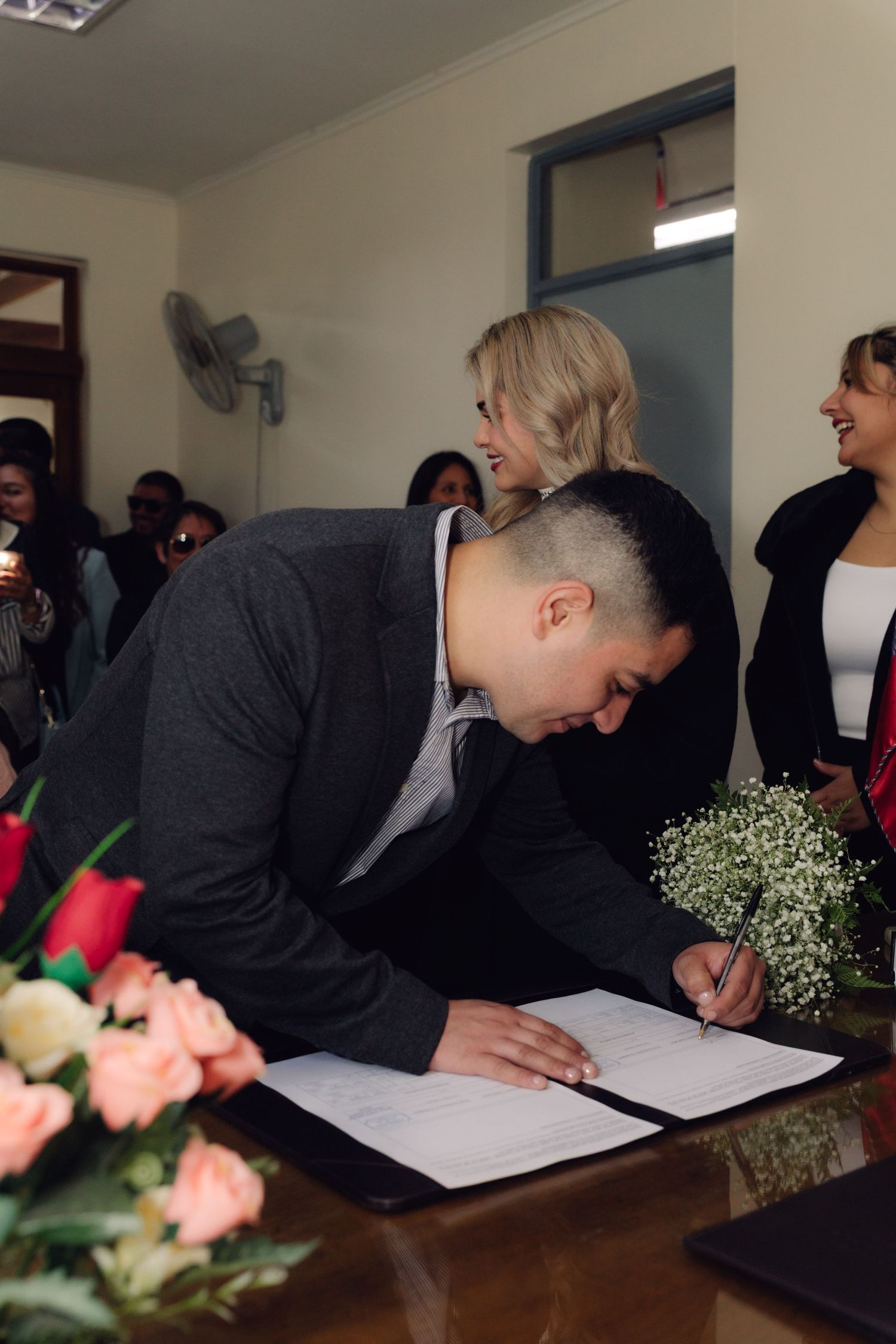 Groom signs official marriage documents at decorated table during civil wedding ceremony with bride nearby.