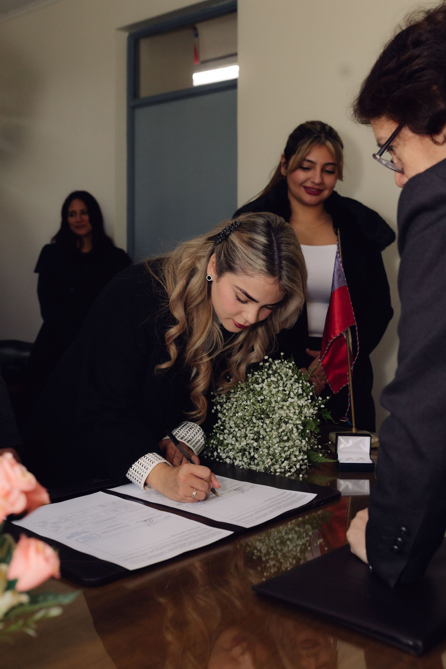 Blonde bride signs official marriage certificate at table while guests and officiant watch during ceremony.