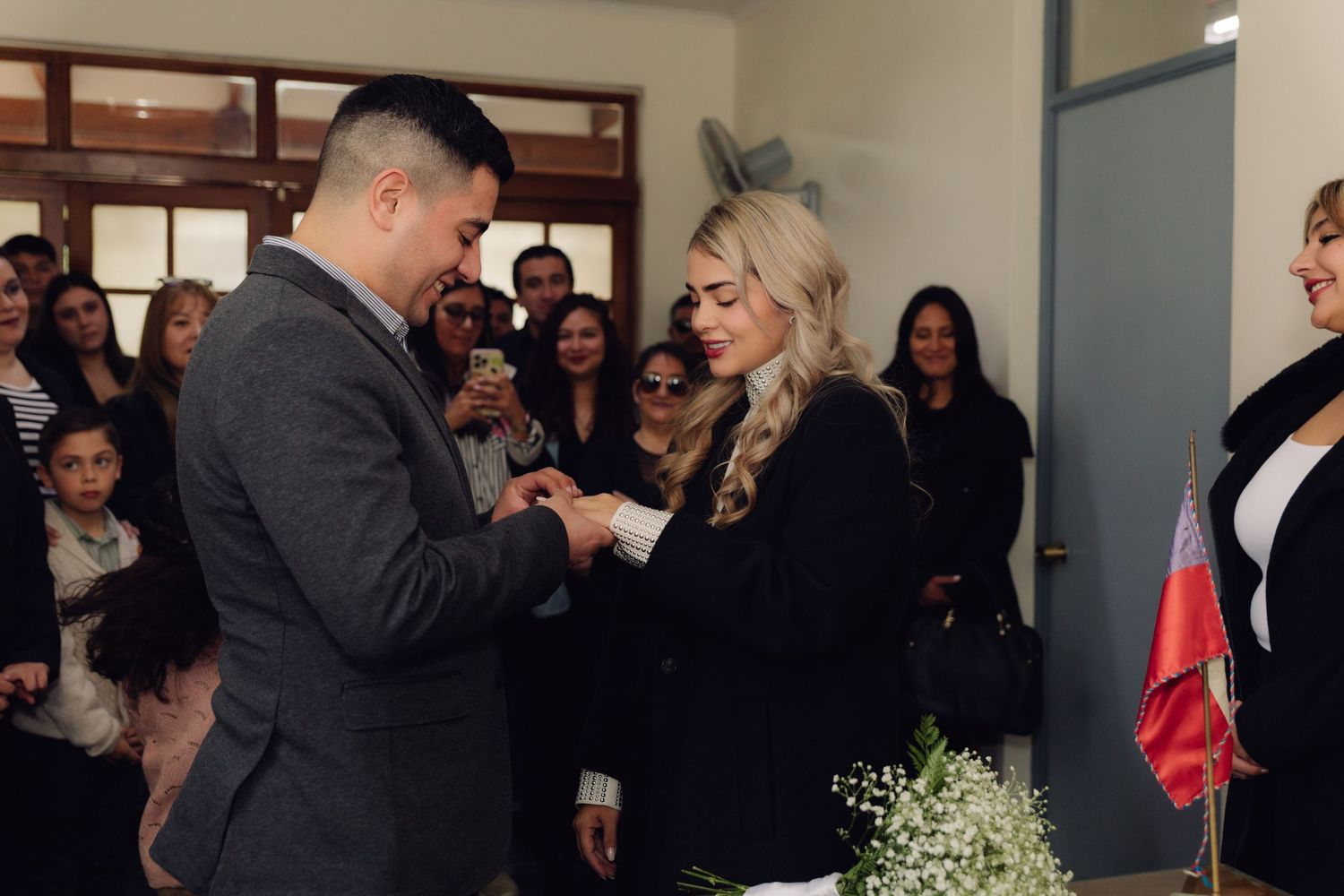 Groom places wedding ring on bride's finger during civil marriage ceremony with guests watching.