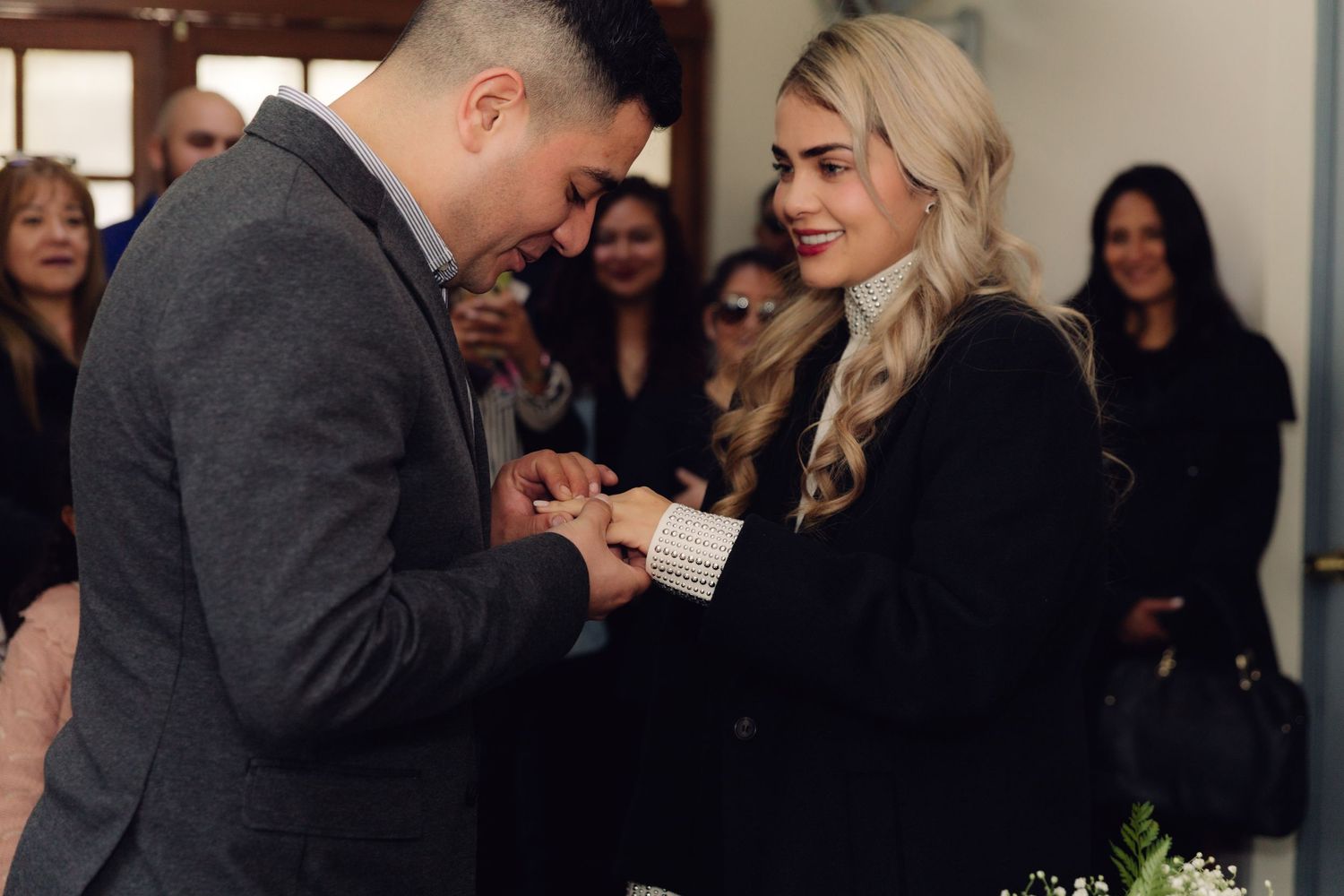 Groom carefully places wedding ring on smiling bride's finger during intimate civil ceremony with guests.