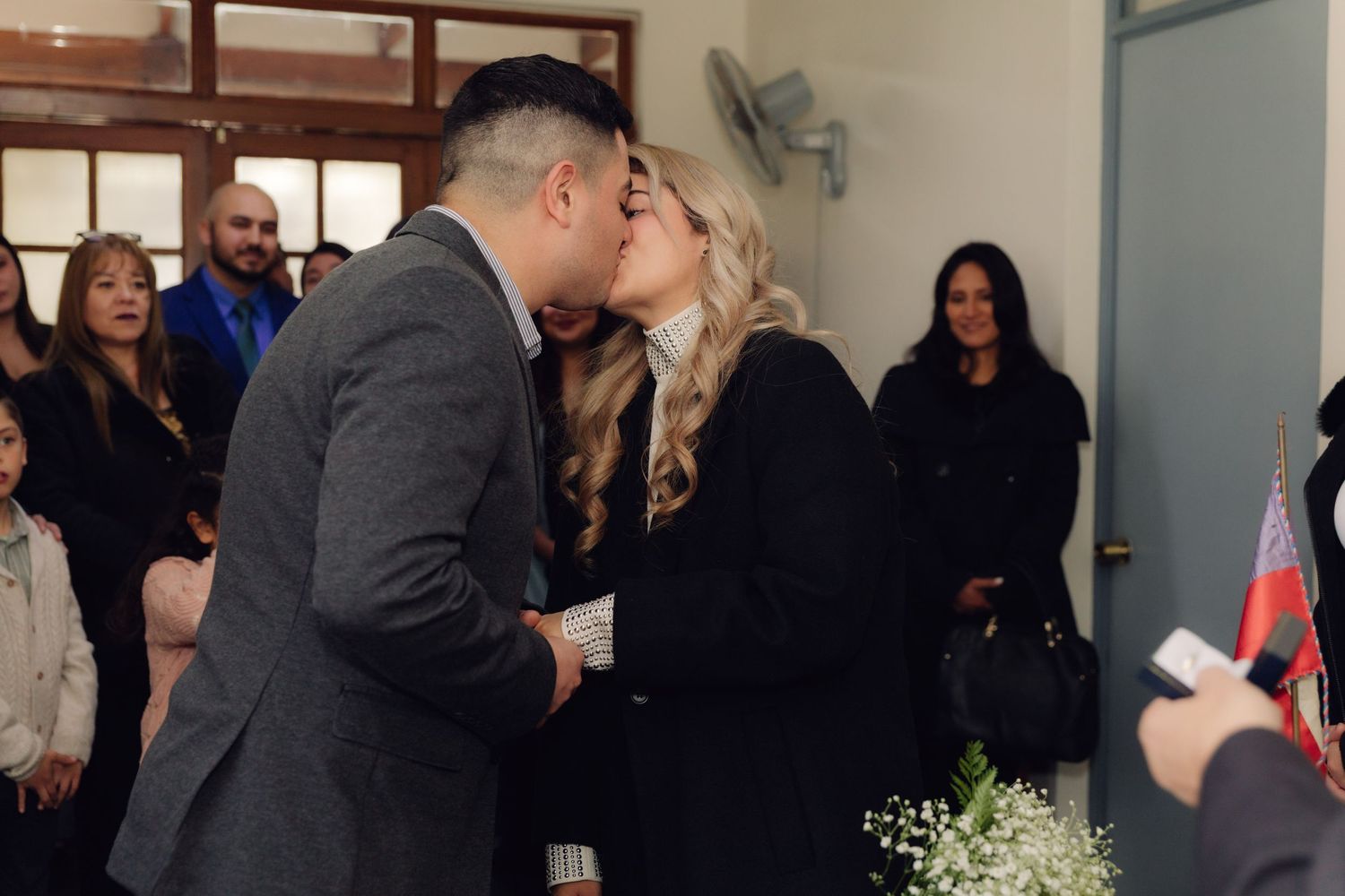 Newlyweds share romantic kiss during civil wedding ceremony as guests watch and celebrate around them.