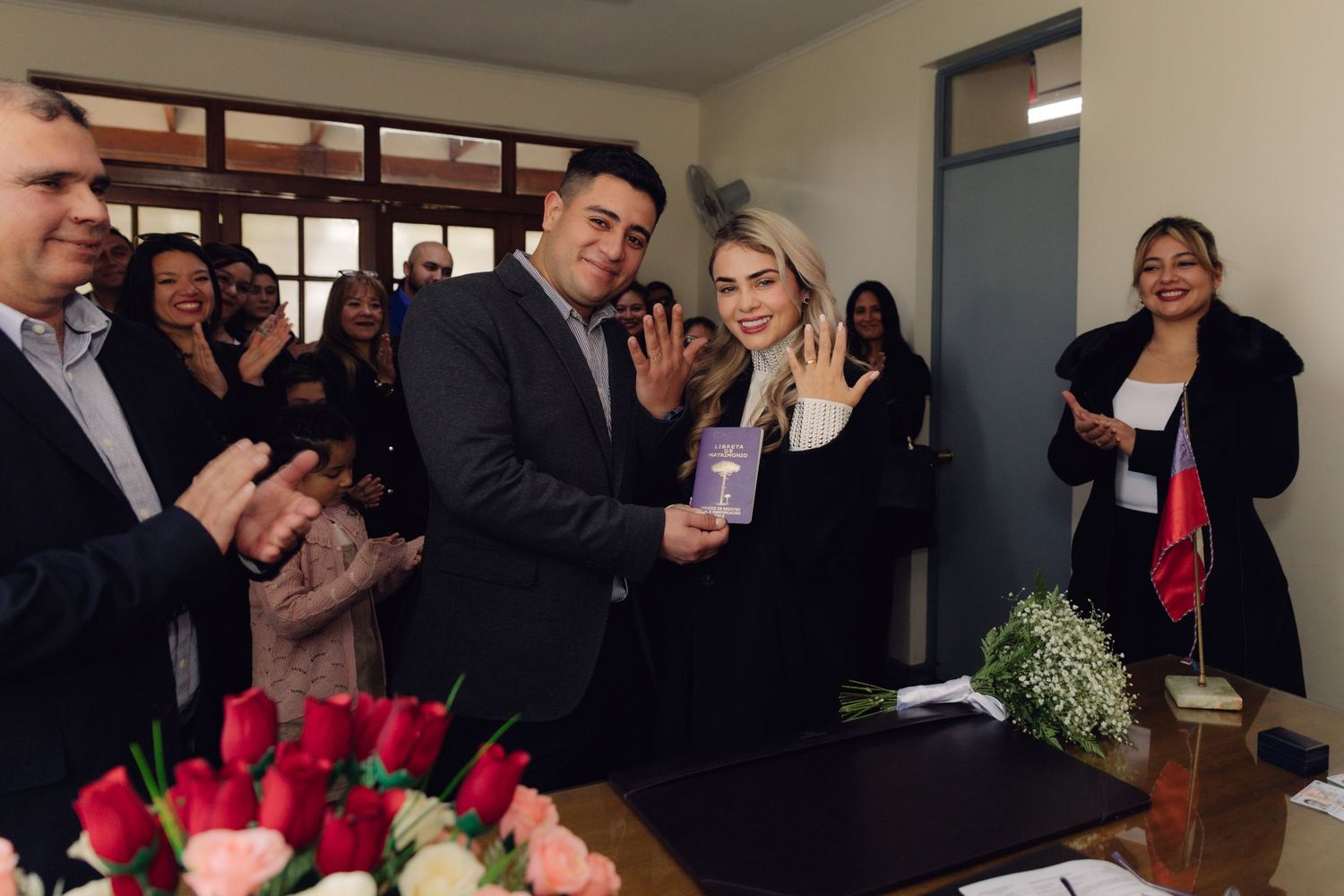Newlyweds pose with marriage booklet as guests applaud beside rose floral arrangements after civil ceremony.