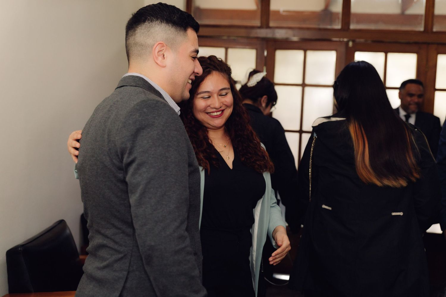 Two people share a warm embrace and smile at a formal gathering indoors.
