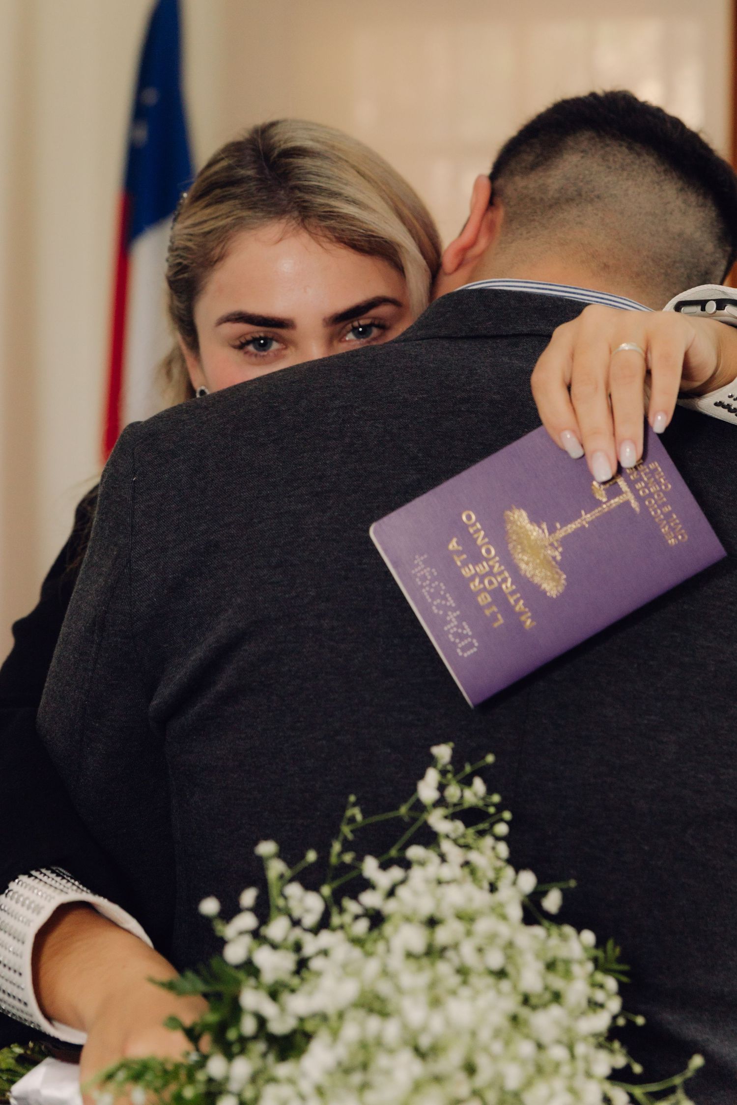 Bride hugs groom tightly, holding marriage certificate and white flower bouquet.