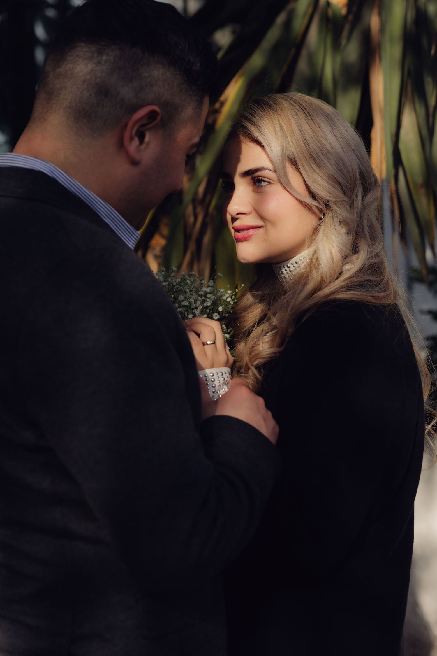 Romantic couple stands close together outdoors near tropical palm foliage.