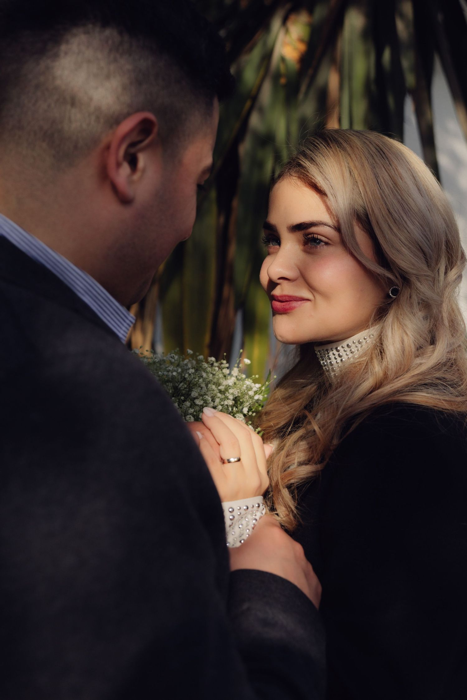 Elegant bride gazes lovingly at groom outdoors near lush green foliage.