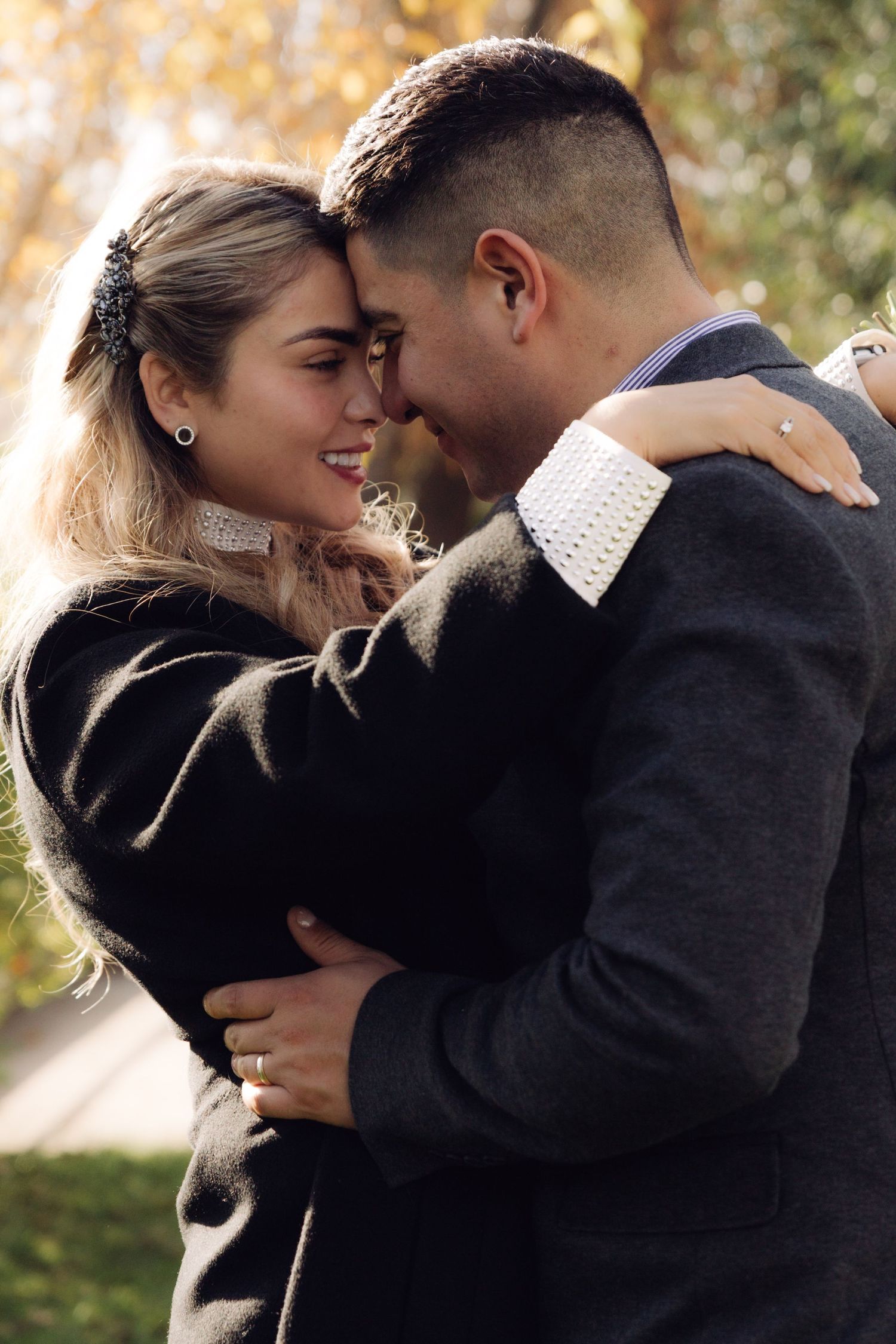 A smiling couple embraces forehead-to-forehead in an autumn park, both wearing dark coats.