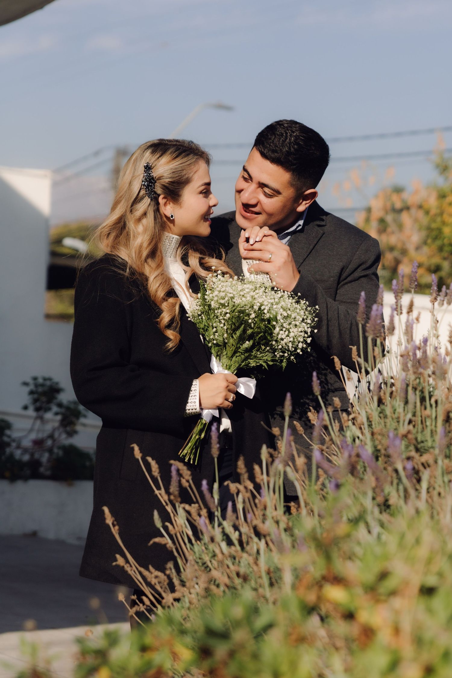 A couple shares a joyful moment outdoors, the woman holding a white wildflower bouquet near lavender plants.