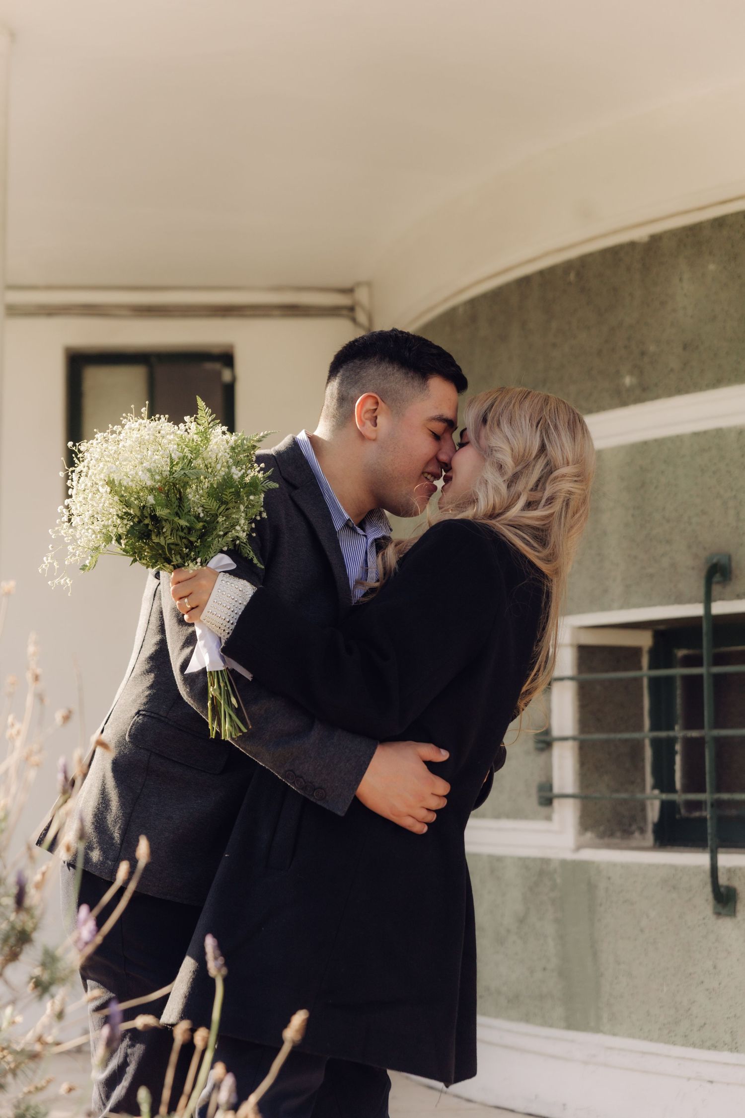 A romantic couple nearly kissing while embracing outdoors, the man holding a white flower bouquet.