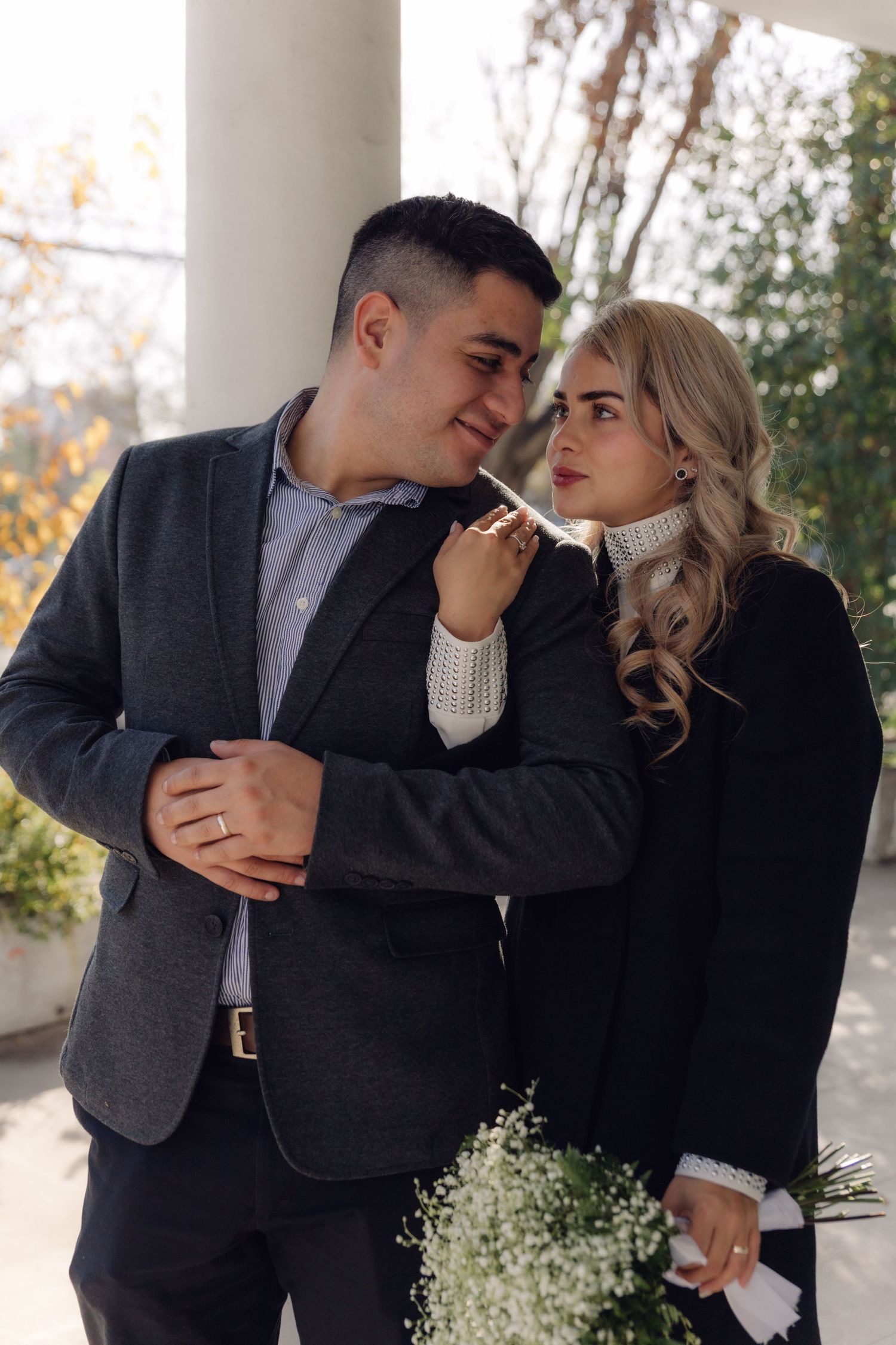 An elegant couple poses under white columns in autumn, the woman touching the mans chin affectionately.
