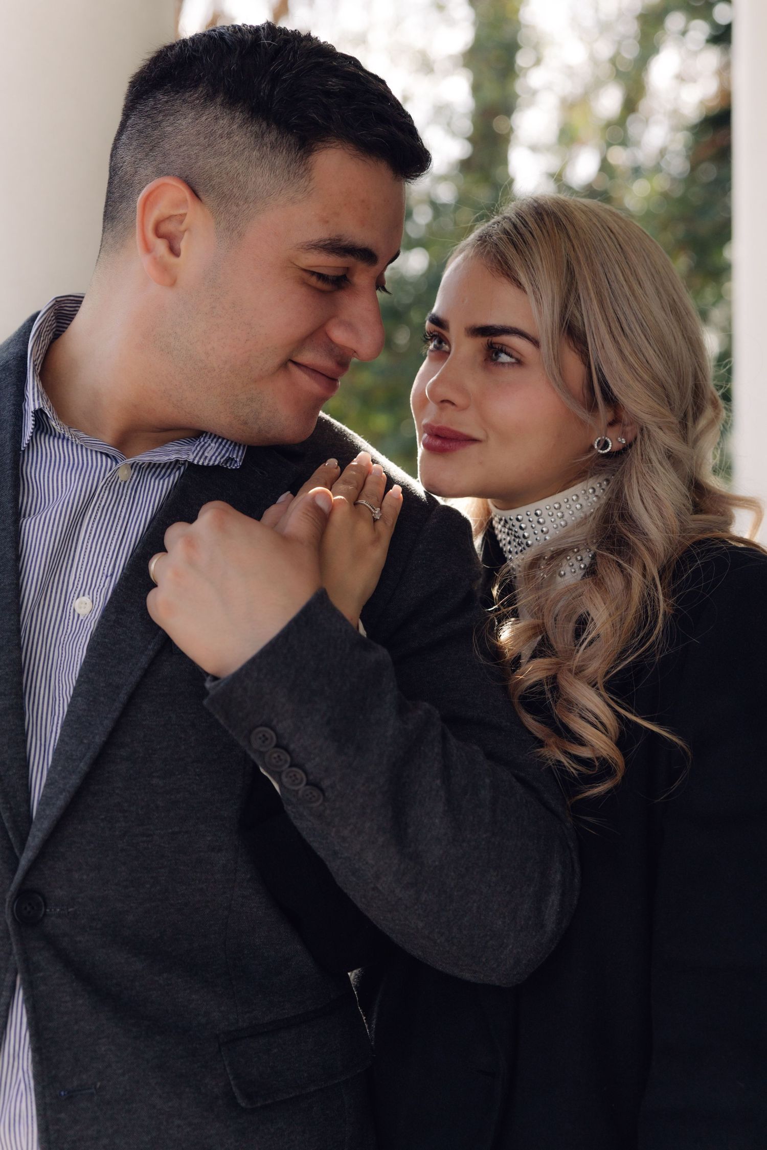 A stylish couple gazes at each other tenderly near white columns, the woman holding the mans lapel.
