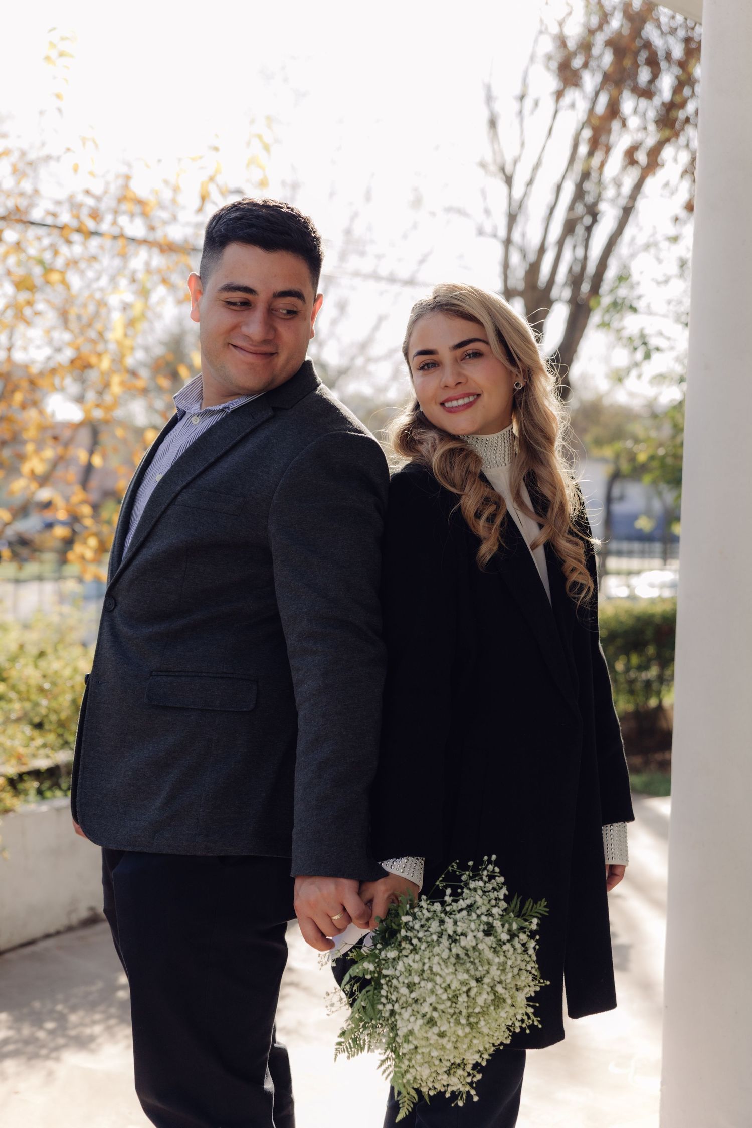 A smiling couple holds hands while posing outdoors in autumn, the woman carrying a white flower bouquet.