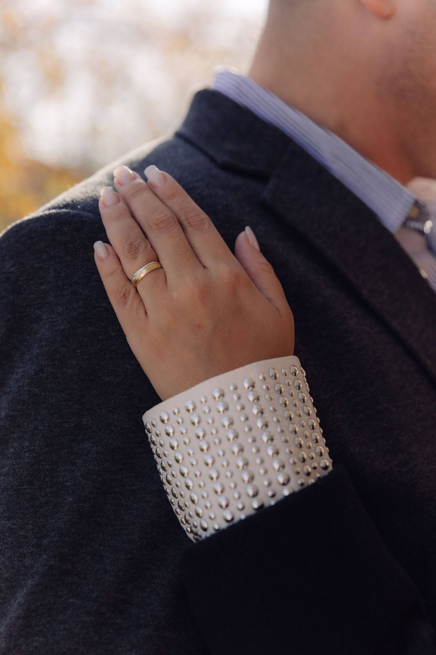A close-up of a womans hand with a gold ring and pearl-studded bracelet resting on a mans dark suit.