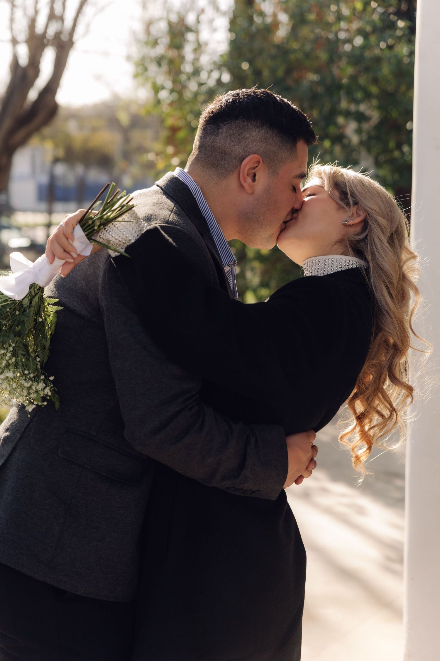 A couple shares a passionate kiss outdoors in sunlight, the man holding a white flower bouquet.
