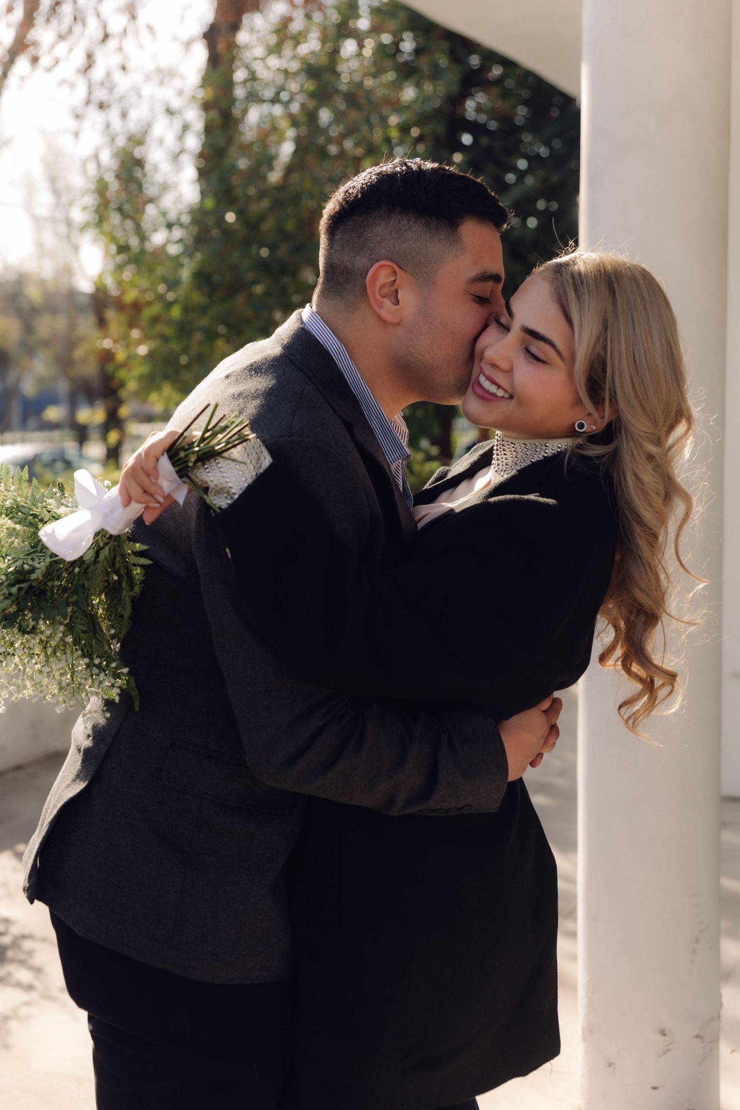 A man kisses a laughing womans cheek while embracing her near white columns, holding a flower bouquet.