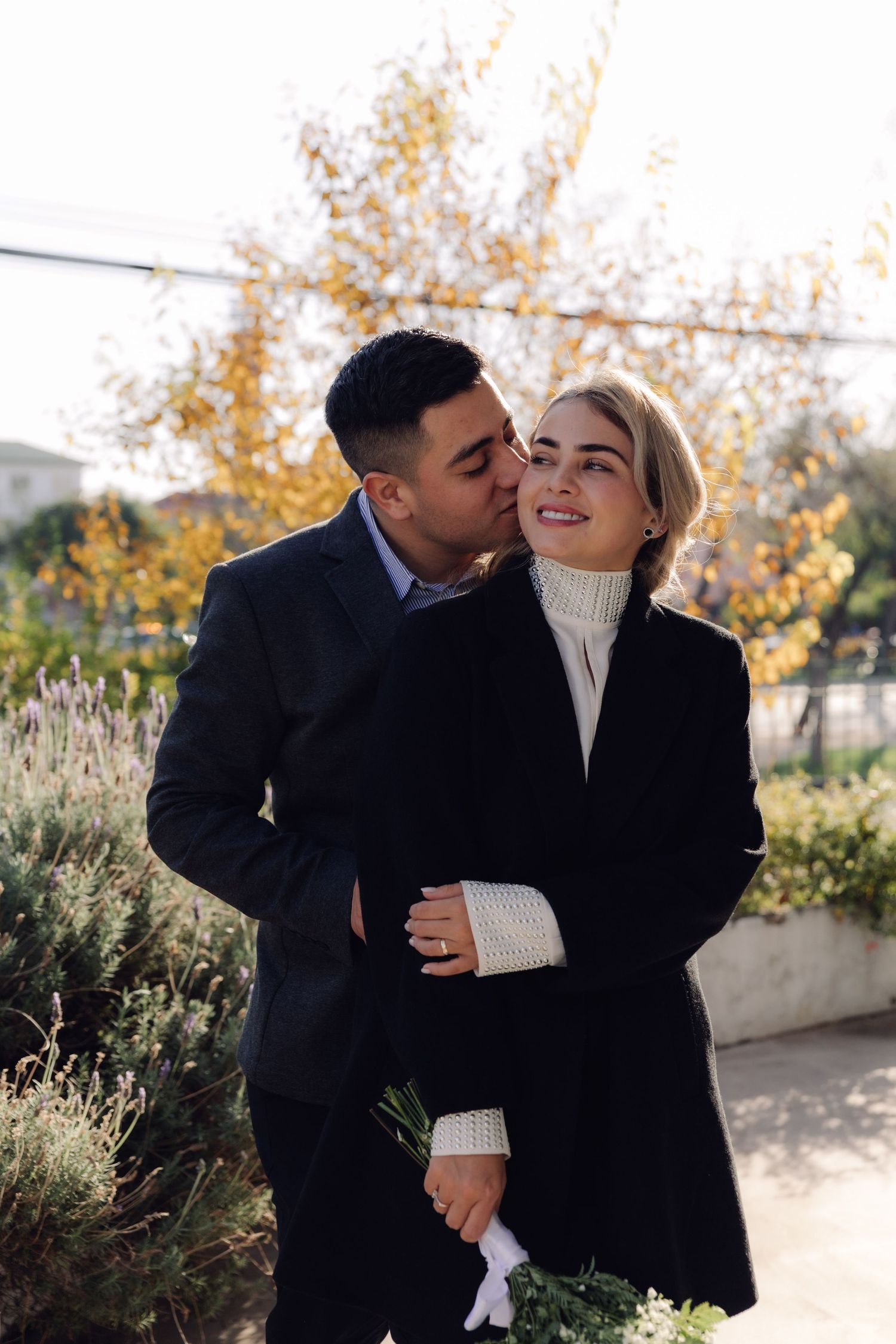A man kisses a smiling womans cheek from behind in a sunny autumn garden, she holds white flowers.