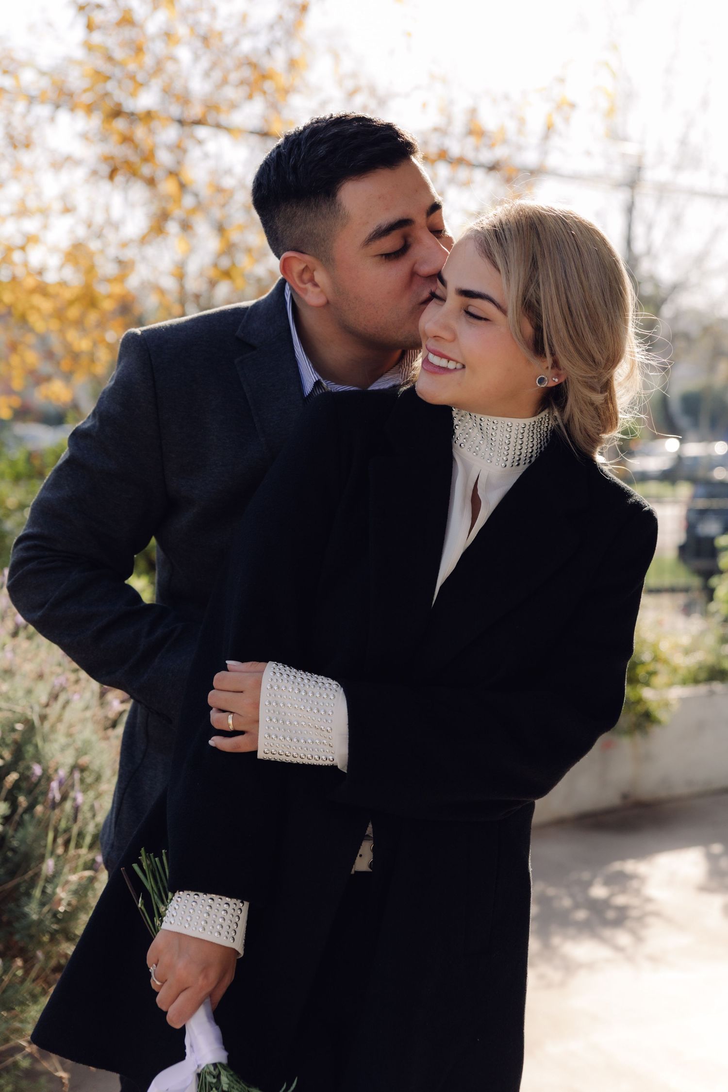 A couple shares a romantic moment outdoors in fall, man kissing woman's cheek as she smiles in black coats.