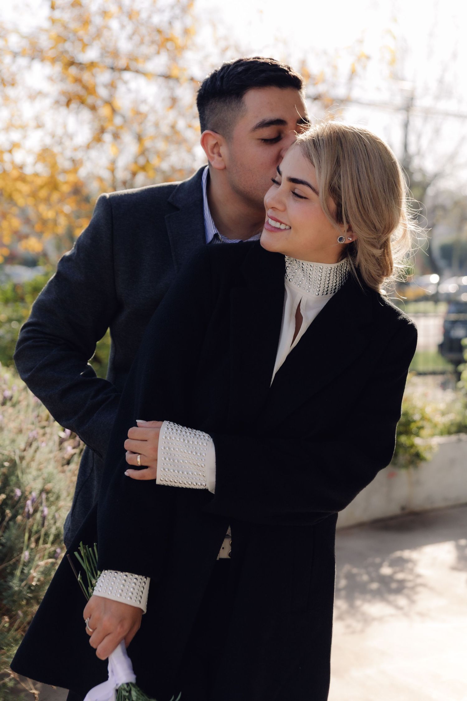 A loving couple embraces outdoors in autumn, man kissing smiling blonde woman's cheek in matching black coats.