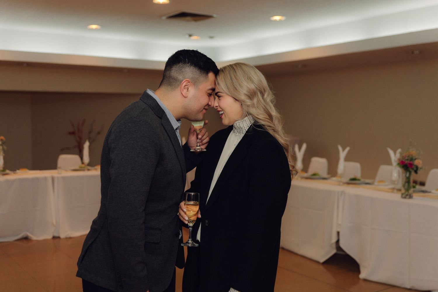 An elegant couple shares an intimate moment at a reception venue, holding champagne glasses and leaning close.
