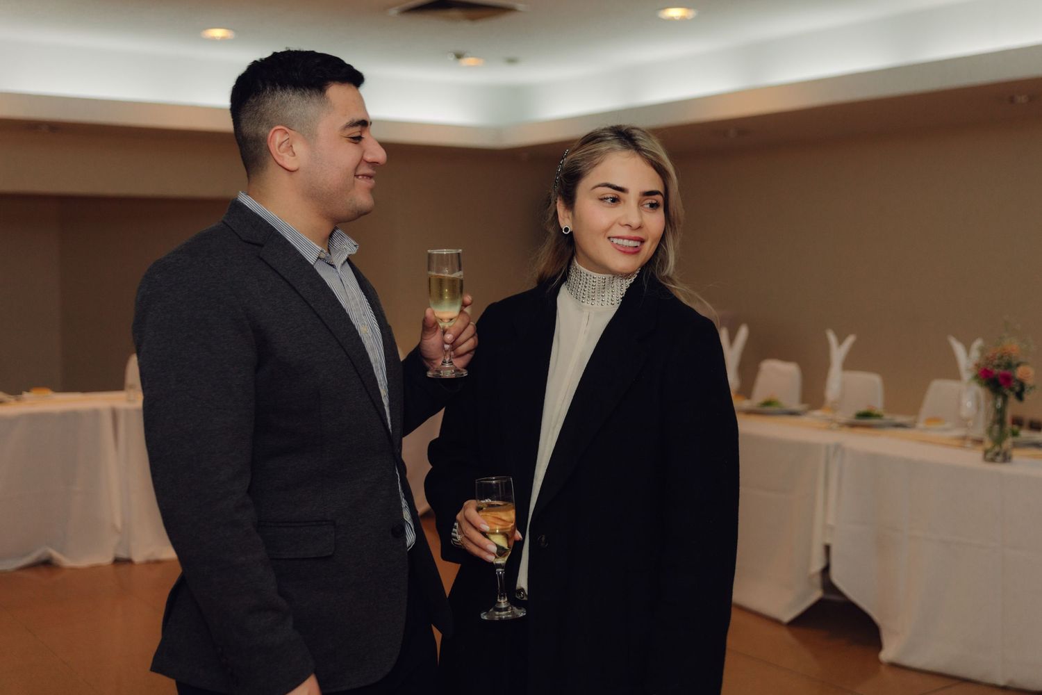 A stylish couple stands together at an indoor event, both holding champagne glasses and smiling warmly.