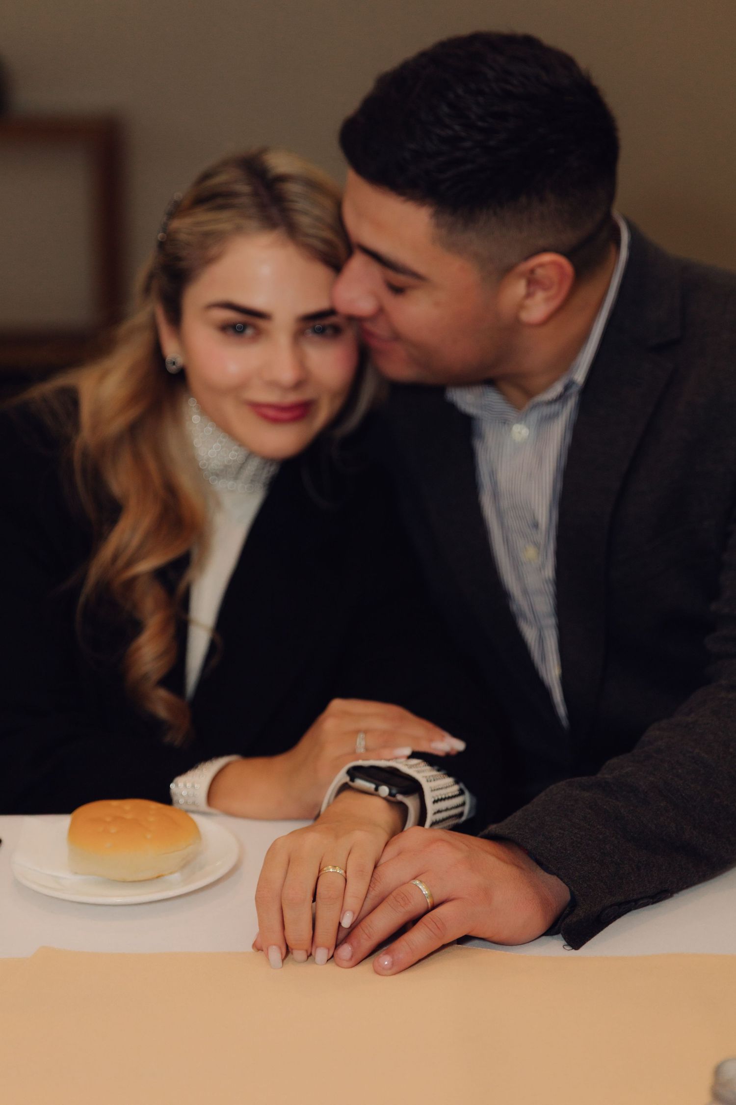 A romantic couple sits at a restaurant table holding hands, man kissing woman's temple as she smiles softly.