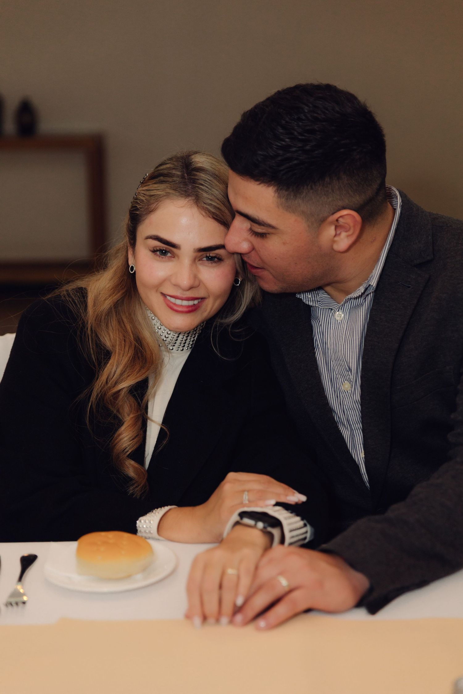 A couple shares a tender moment at a restaurant, man kissing smiling woman's temple while holding hands at table.